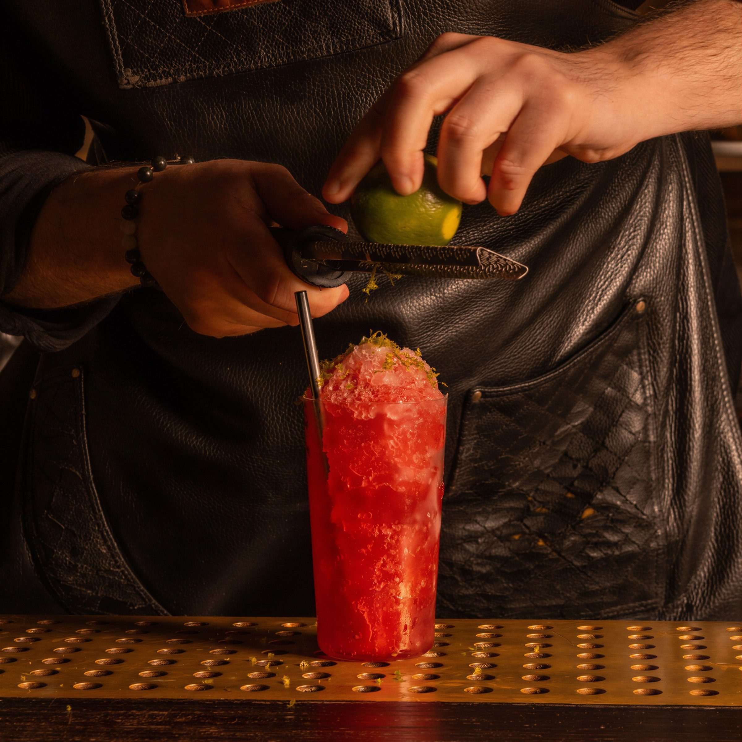 Hands grating lime over a red iced drink with a metal straw. Person wearing a black leather apron. Warm, dimly lit bar setting.
