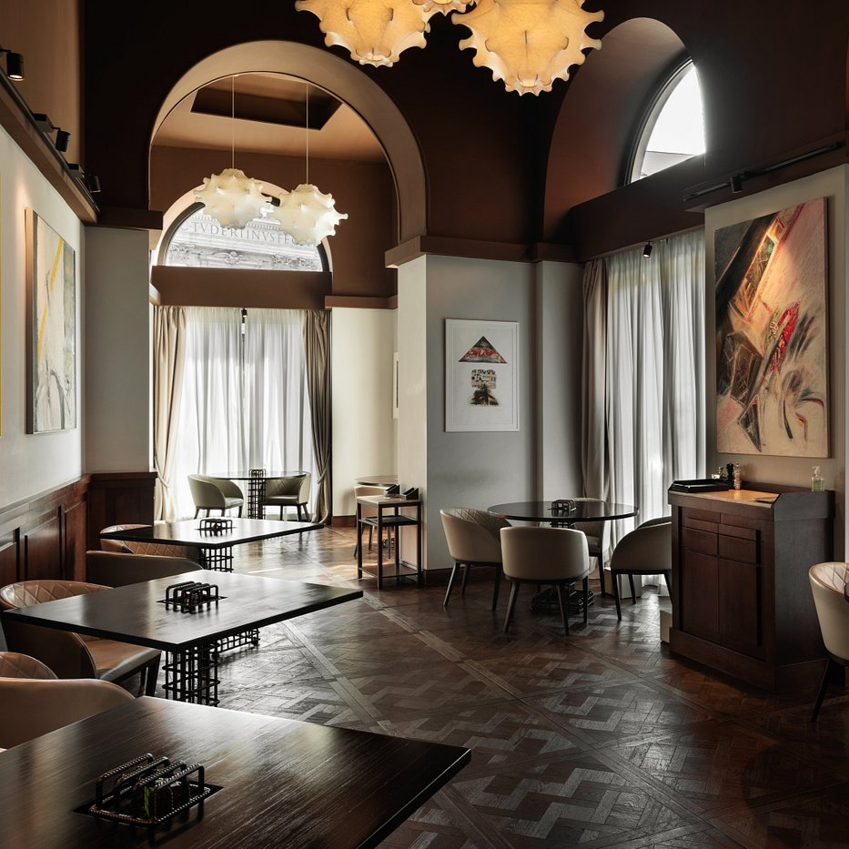 Elegant restaurant interior with arched windows, abstract art, and modern pendant lights. Round tables and beige chairs on patterned floor. Cozy ambiance.