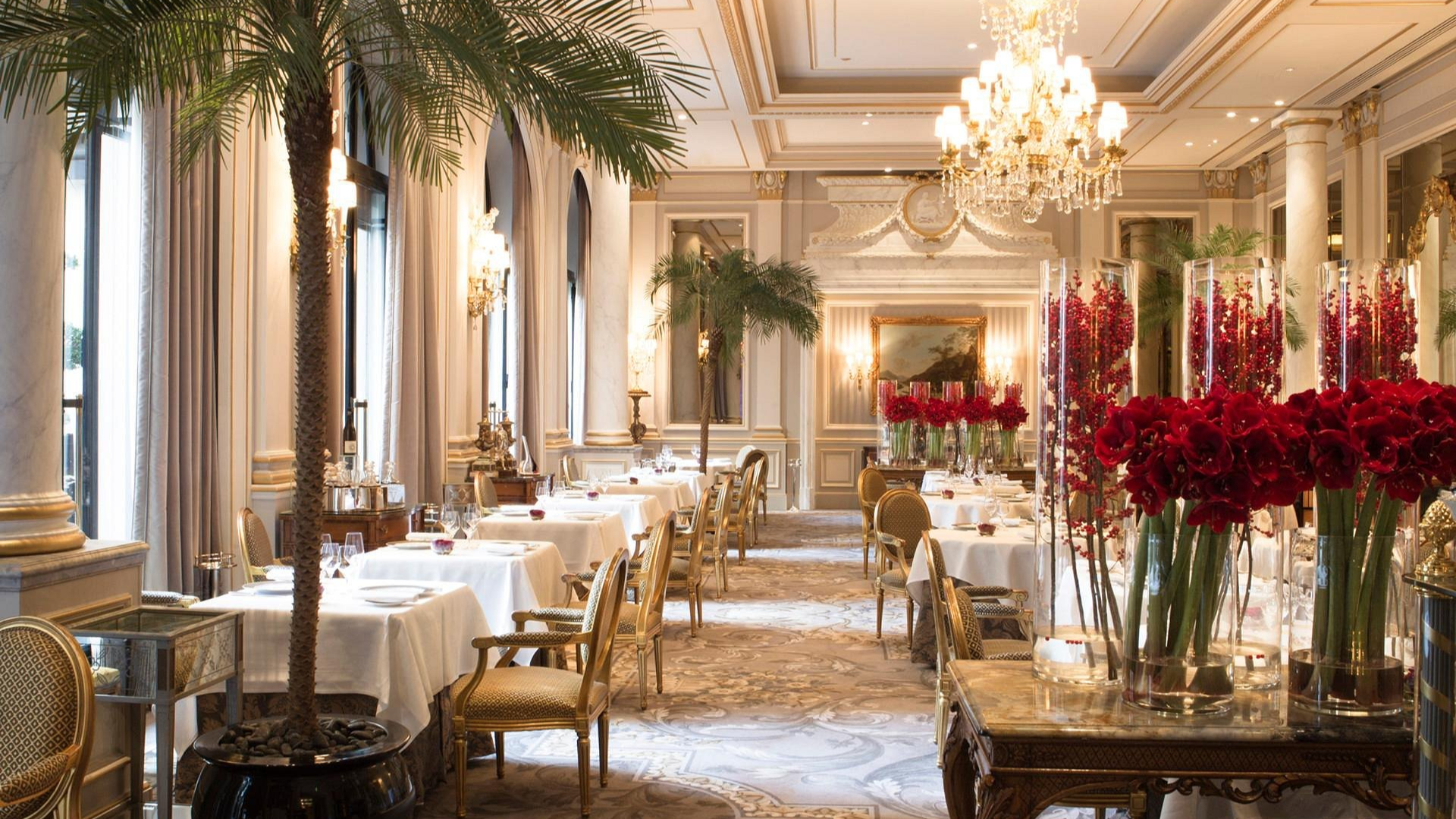 Elegant restaurant interior, featuring white tablecloths, red floral arrangements, chandeliers, and palm trees; a luxurious and serene ambiance.