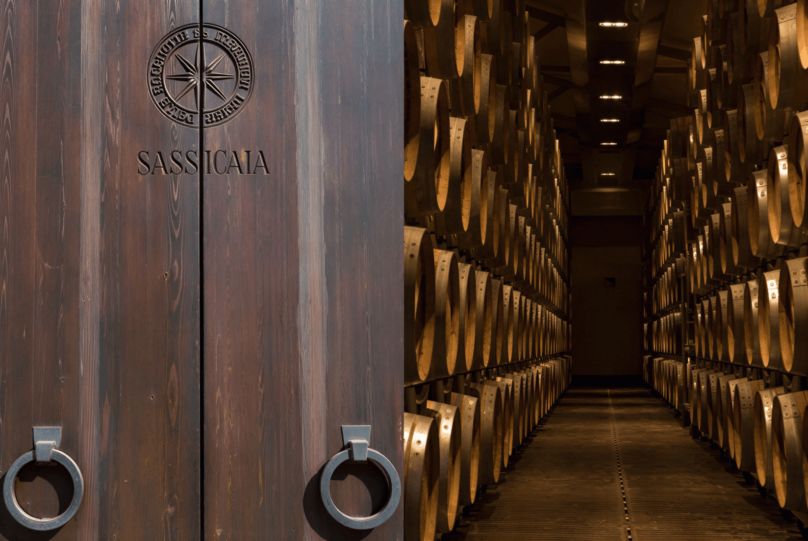 Wooden cellar door of Sassicaia Barrel Room at Tenuta San Guid, flanked by two handles, opens to reveal rows of wine barrels in a dimly lit storage room.