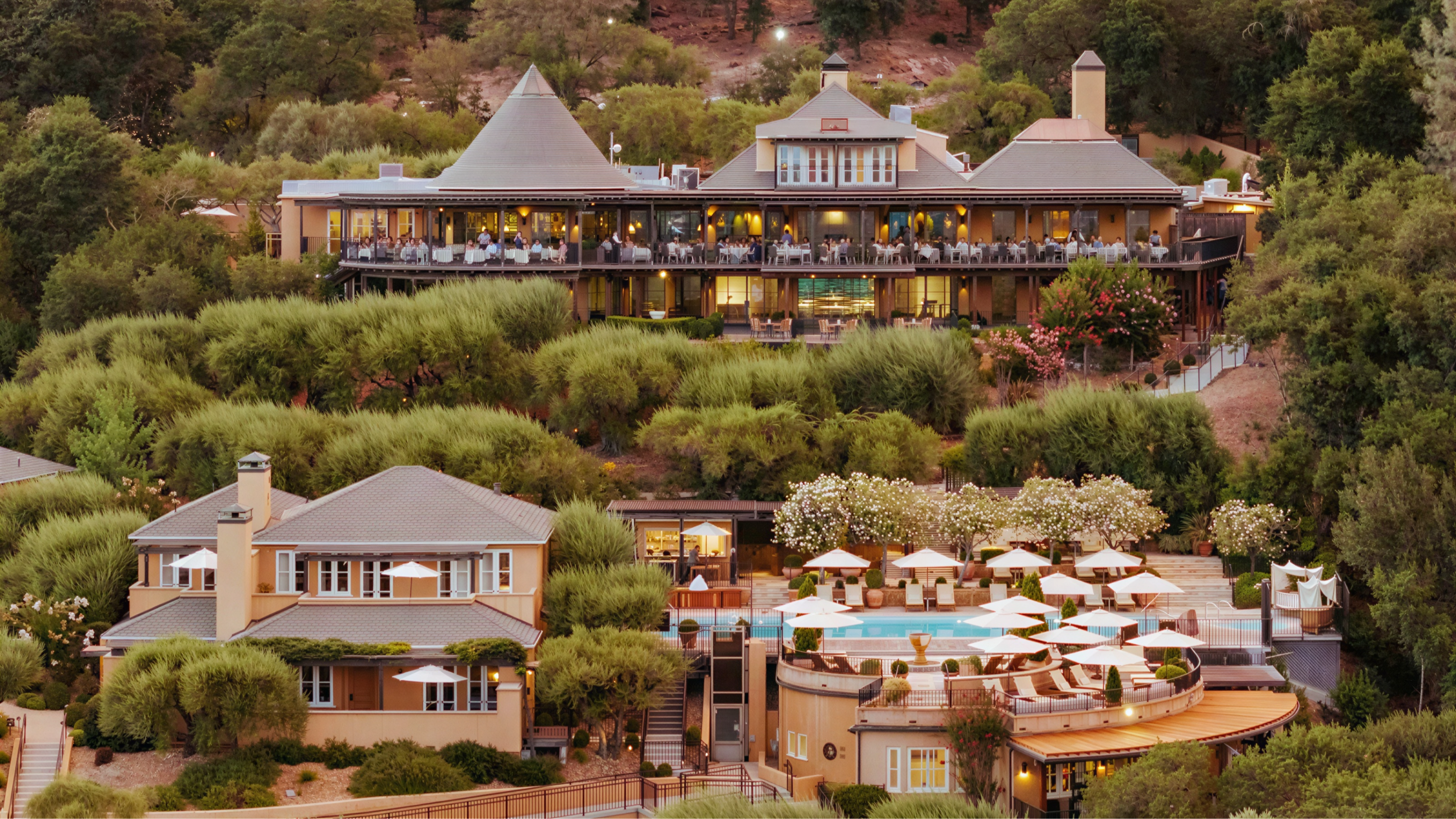 Luxurious hillside resort with terraced gardens, two-story buildings, umbrellas, and a pool. Surrounded by lush greenery. Relaxed atmosphere.