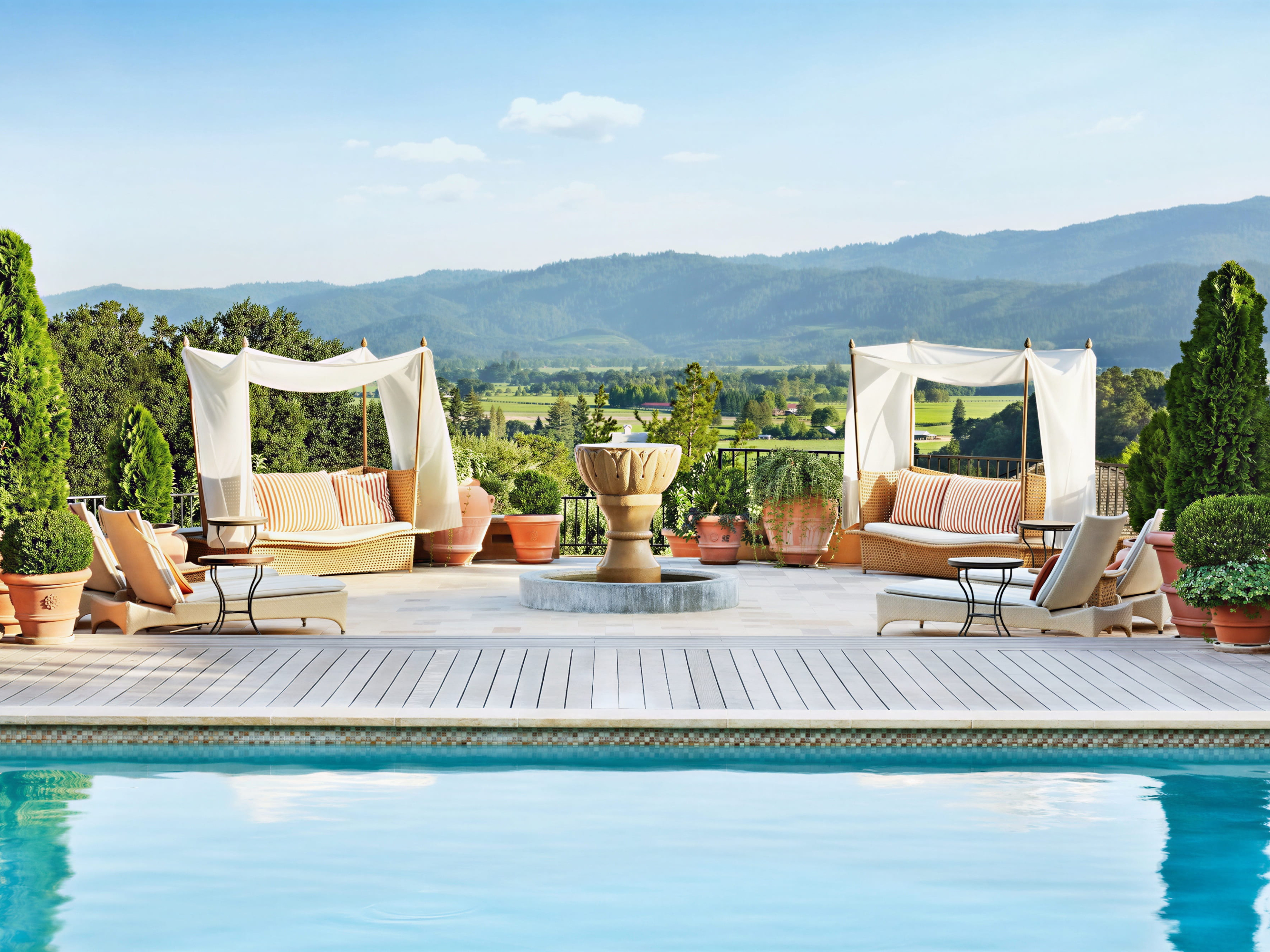 Elegant poolside scene with loungers, canopied sofas, and a fountain. Lush greenery and mountains in the background enhance tranquility.