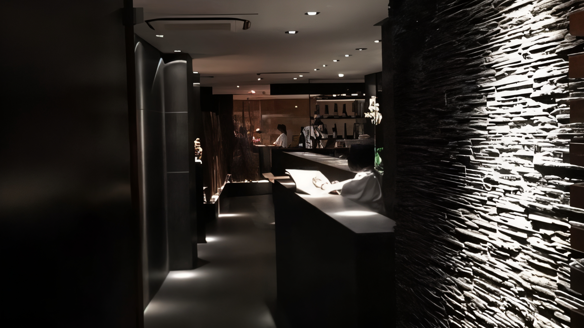 Dimly lit modern hotel lobby with textured walls. Staff at the reception desk; ambient lighting creates a sleek, calm atmosphere.