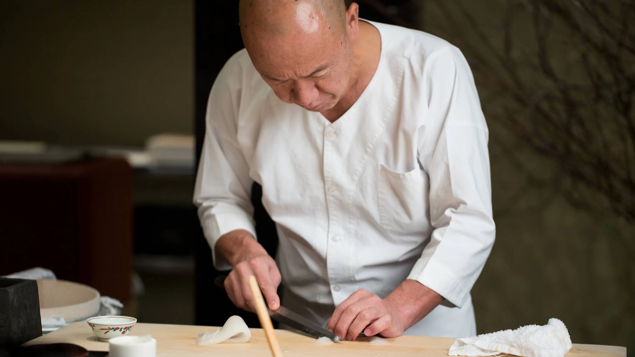 Chef in a white coat skillfully slices food on a wooden board in a dimly lit kitchen. Sparse setup with a focused, calm atmosphere.