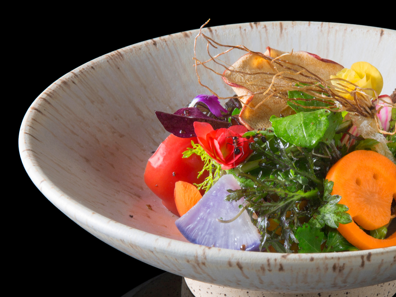 A white ceramic bowl holds a colorful salad with sliced carrots, radishes, leafy greens, and edible flowers against a black background.