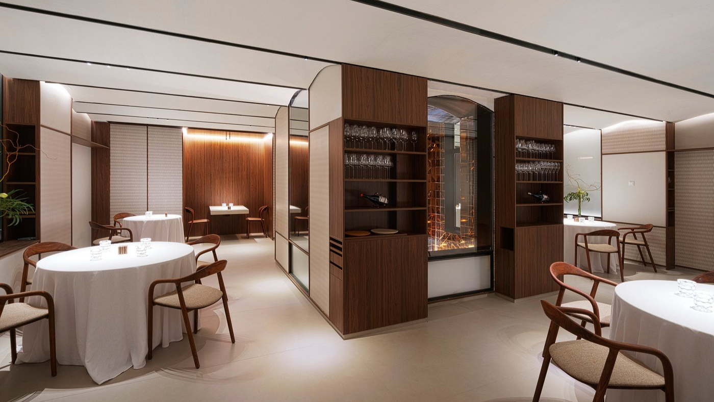 Elegant restaurant with white tablecloths and wooden chairs, warm lighting, and shelves with glasses. Calm, modern ambiance.