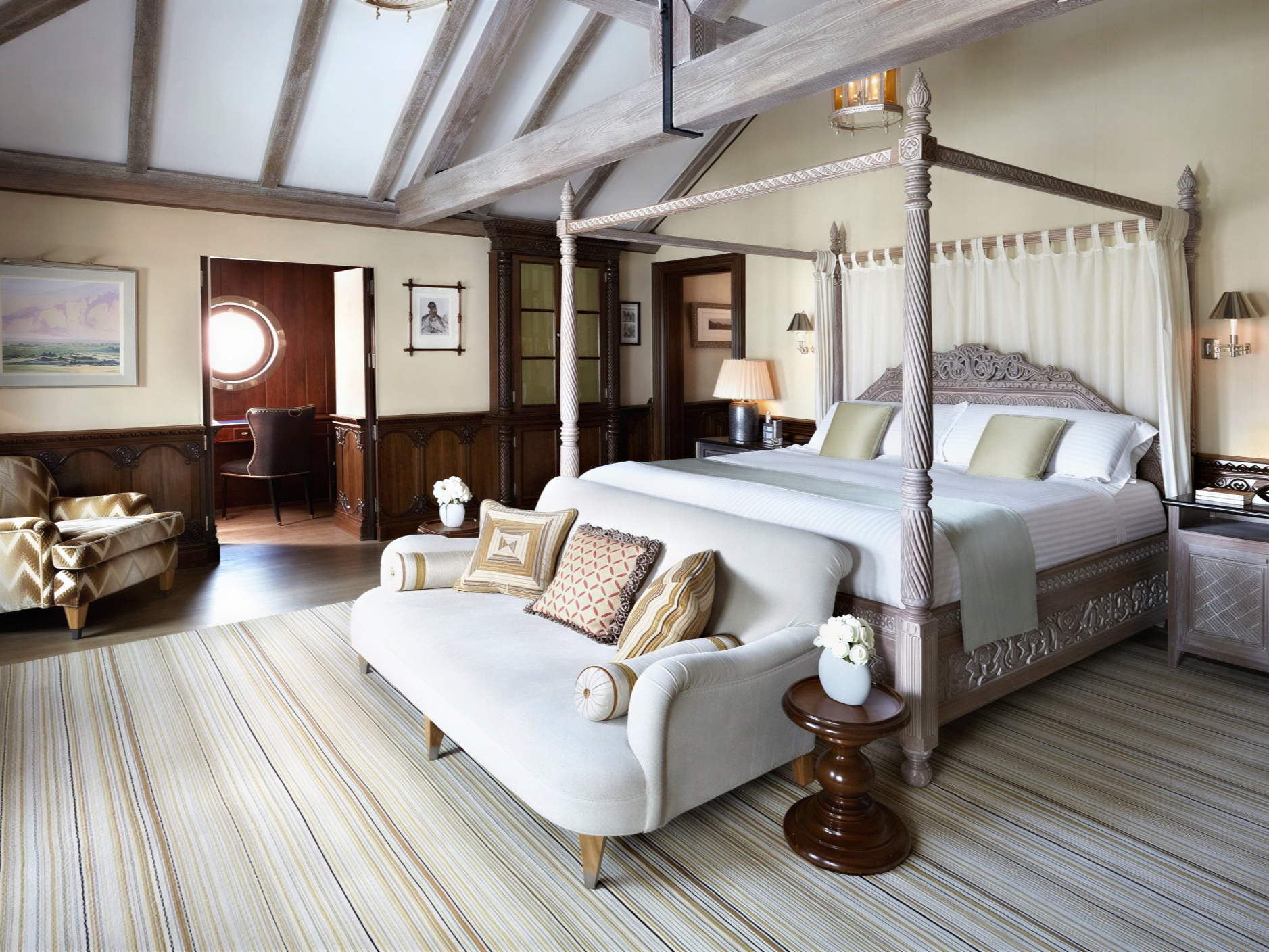 Elegant bedroom with a carved four-poster bed, striped rug, and a cozy white sofa. Soft lighting and a porthole window create a serene mood.