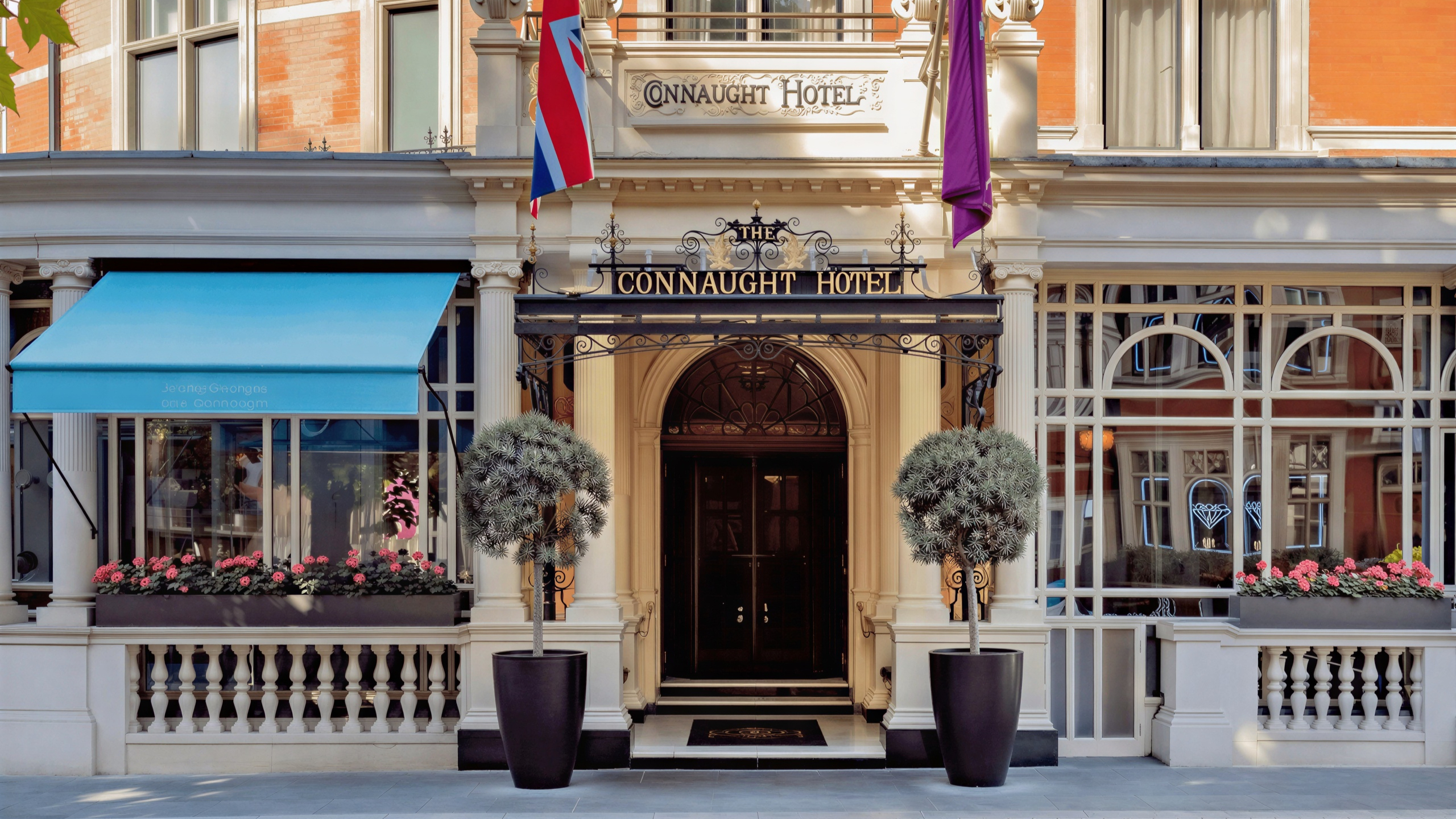 Elegant hotel entrance with "Connaught Hotel" sign, flanked by two potted trees. Blue awning and UK flag present. Flower boxes below windows.