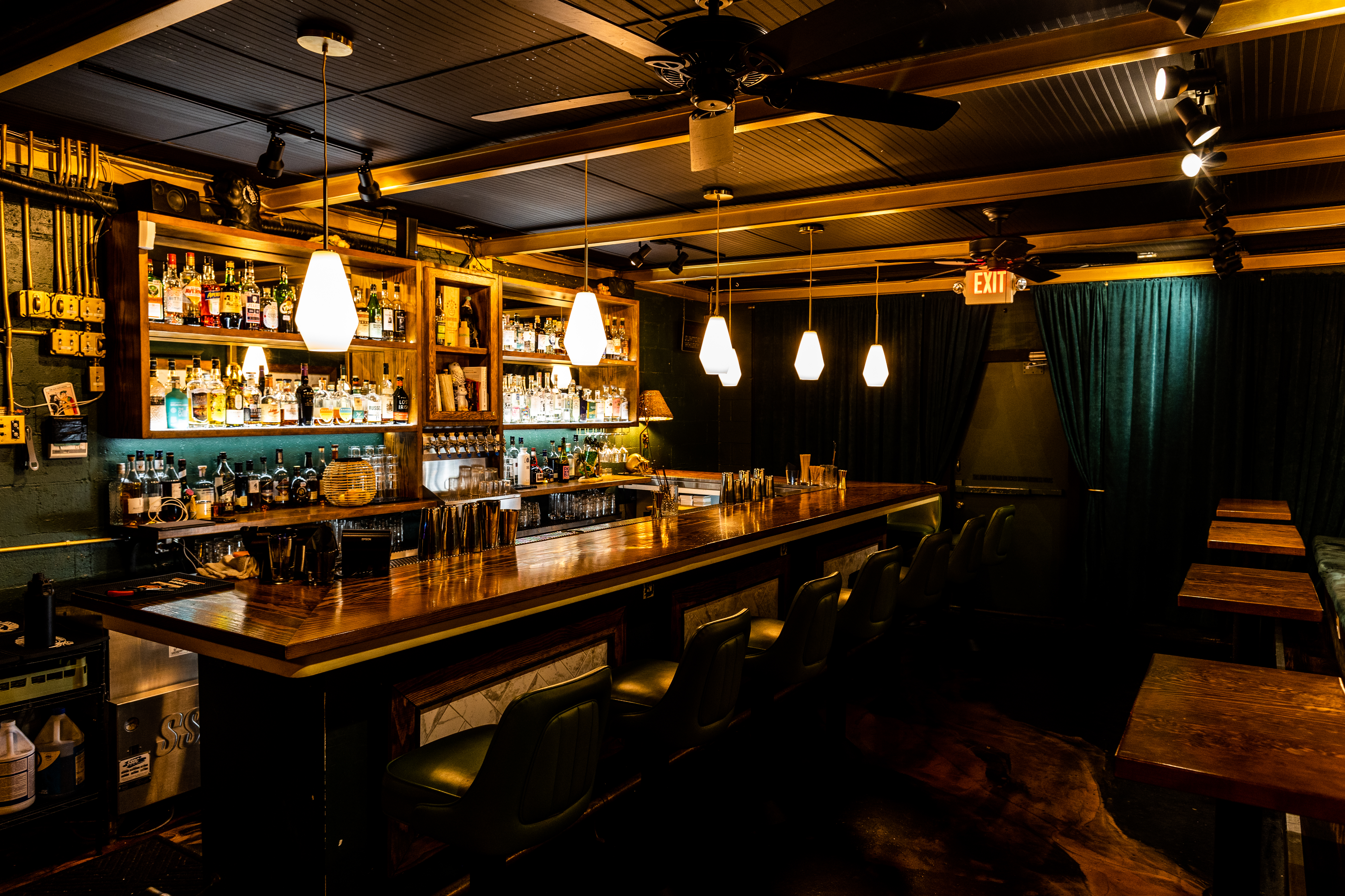 Cozy bar with wooden counter, shelves stocked with bottles. Pendant lights cast a warm glow. Green velvet curtains and an exit sign visible.