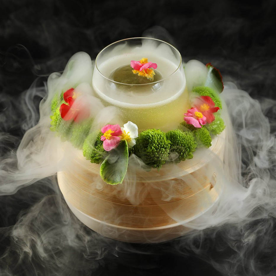 A drink in a glass sits on a wooden base, surrounded by colorful flowers and green moss, with mist enveloping it against a dark background.