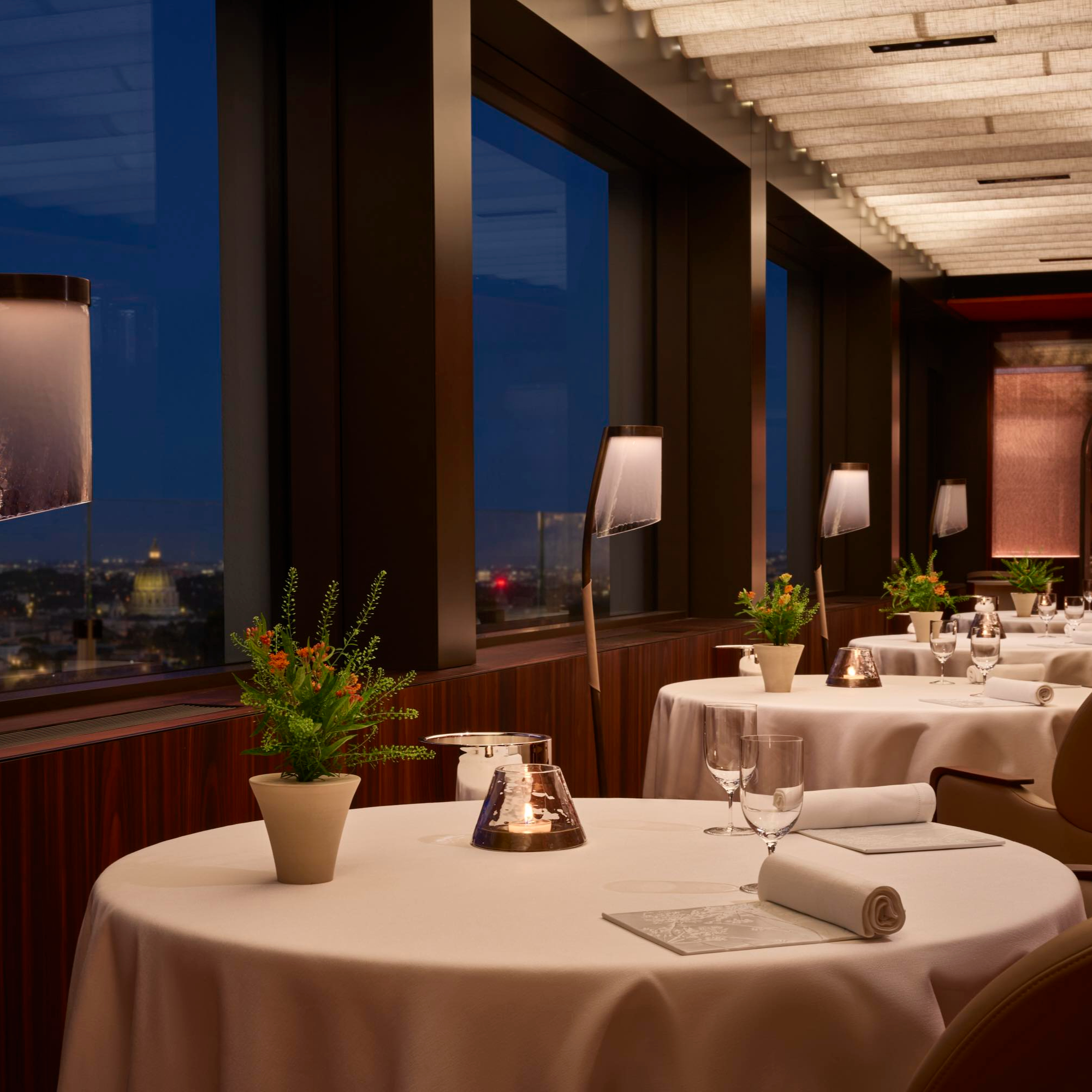 Elegant restaurant interior with neatly set round tables, white tablecloths, glassware, and potted plants, overlooking a nighttime cityscape.