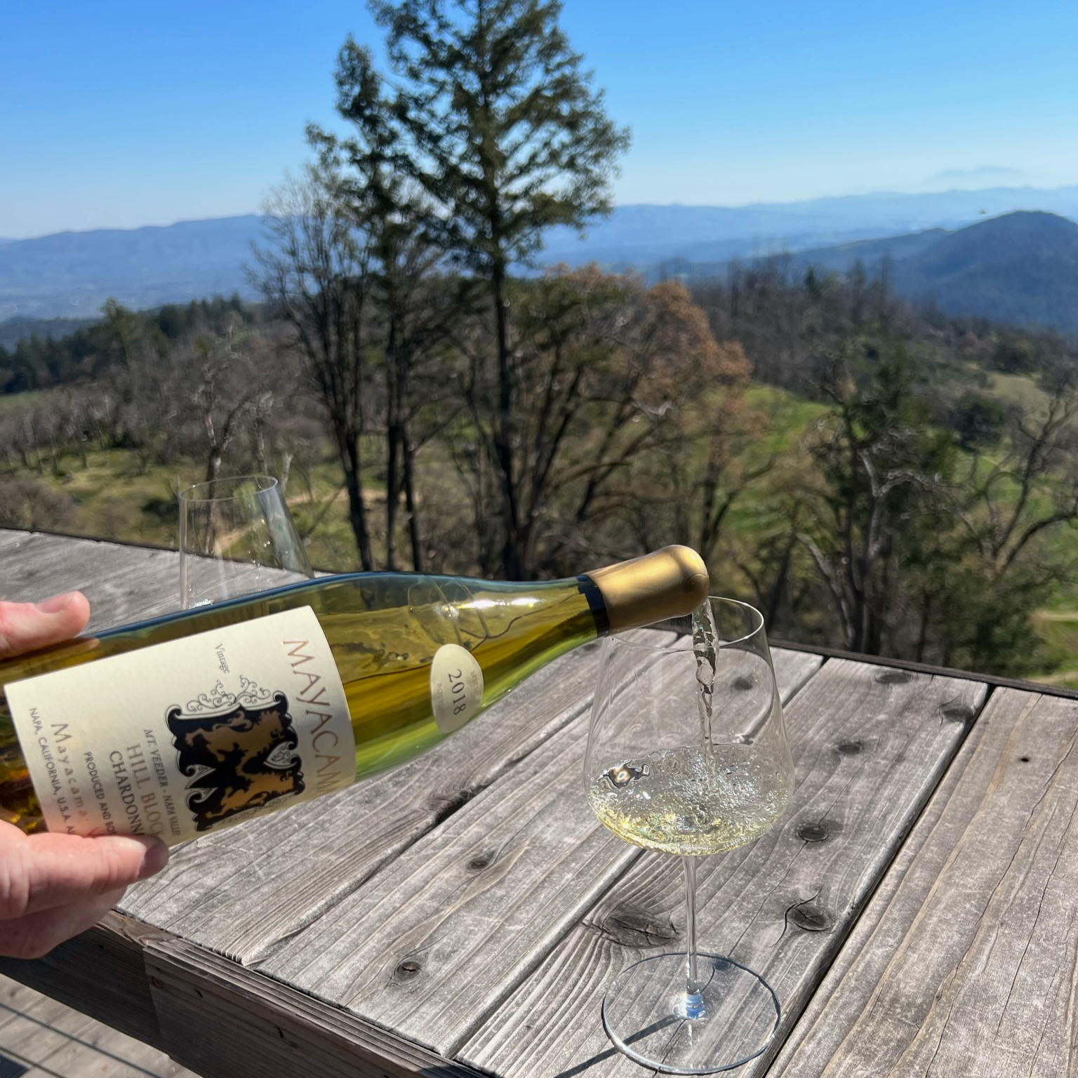 Wine being poured into a glass on a wooden table, overlooking a scenic landscape with distant mountains under a clear blue sky.