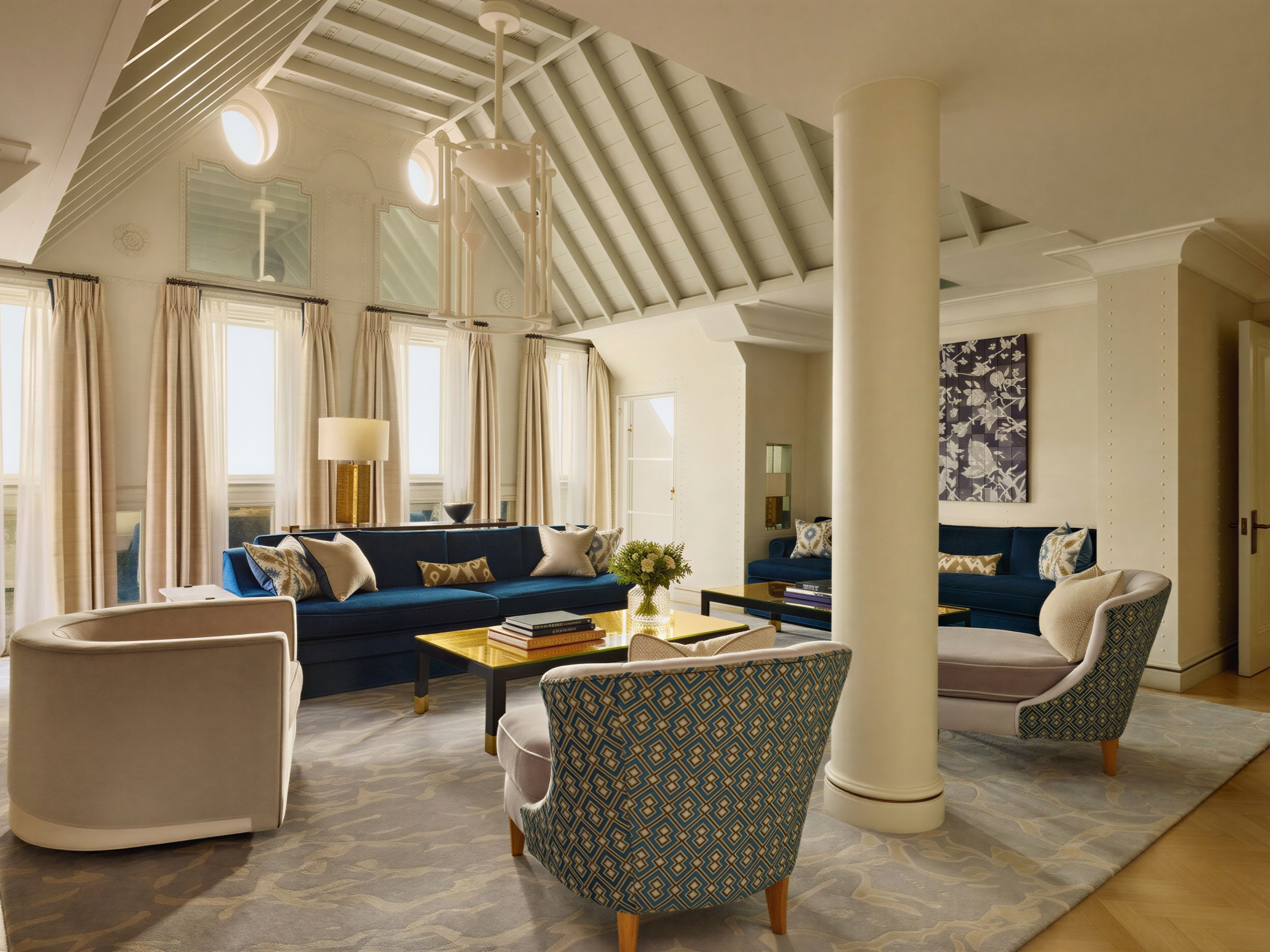 Spacious living room with blue sofas, patterned chairs, and large windows. Beige and blue decor. A pillar and floral artwork are visible.