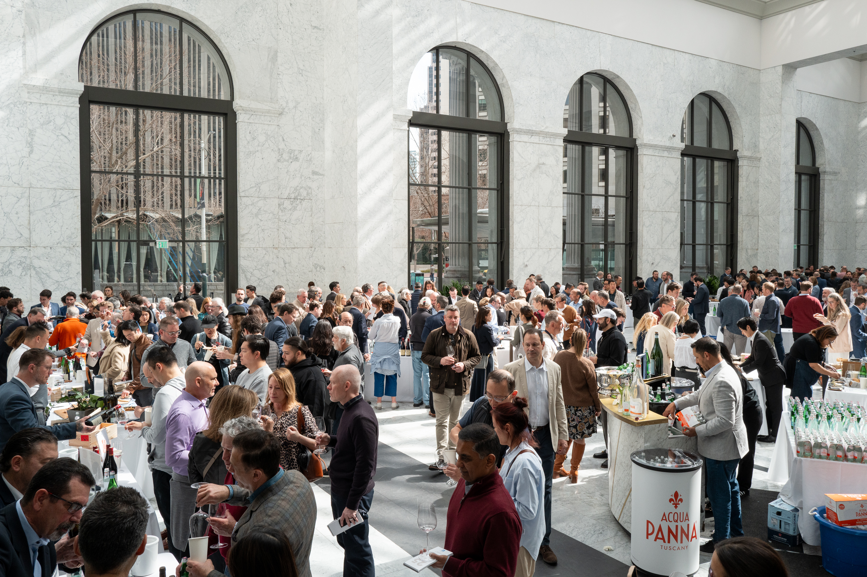 Crowded indoor event with people mingling around tables, some holding drinks. Large windows, marble walls, and "Acqua Panna" visible. Bright ambiance.