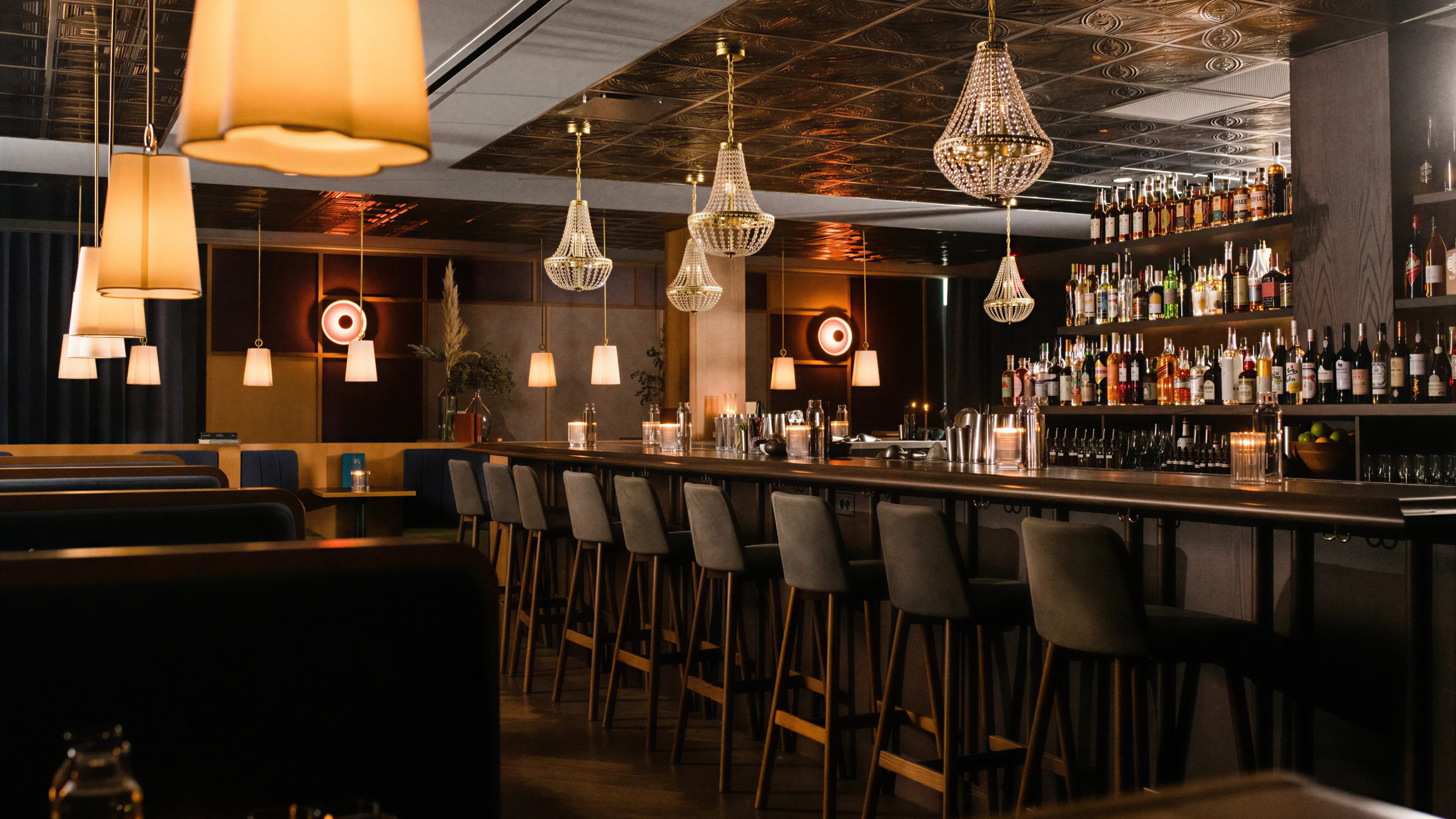 Chic bar interior with empty stools, pendant lights, crystal chandeliers, and shelves stocked with bottles. Warm, inviting ambiance.