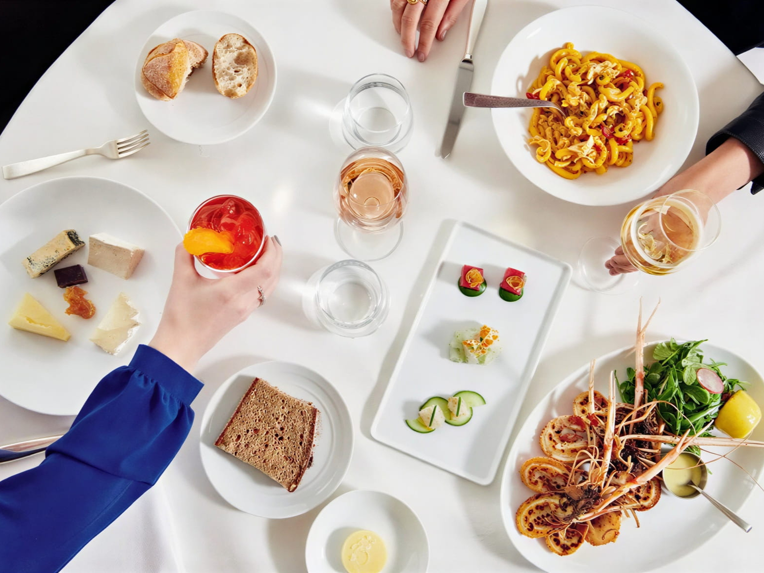 Aerial view of a selection of dishes from Marea NYC like pasta, cheese, and shrimp. People holding drinks, set on a white tablecloth.