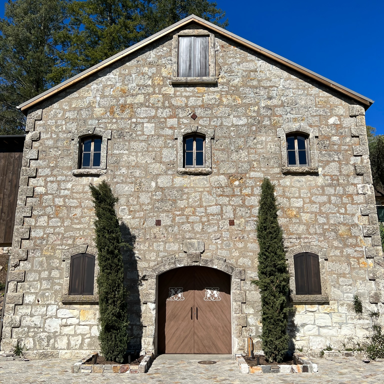 Stone building with arched wooden doors, two tall trees flanking entrance. Blue sky background, rustic and tranquil setting.