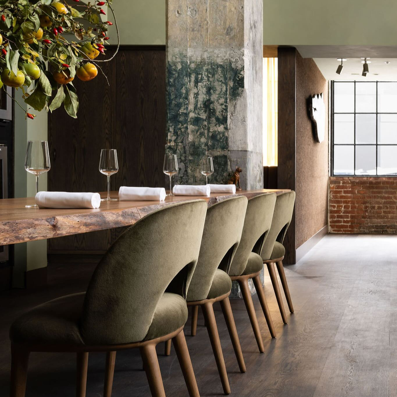 Dining room with green chairs, rustic table set with glasses, and napkins. Fruit decor hangs overhead. Exposed brick and window in background.