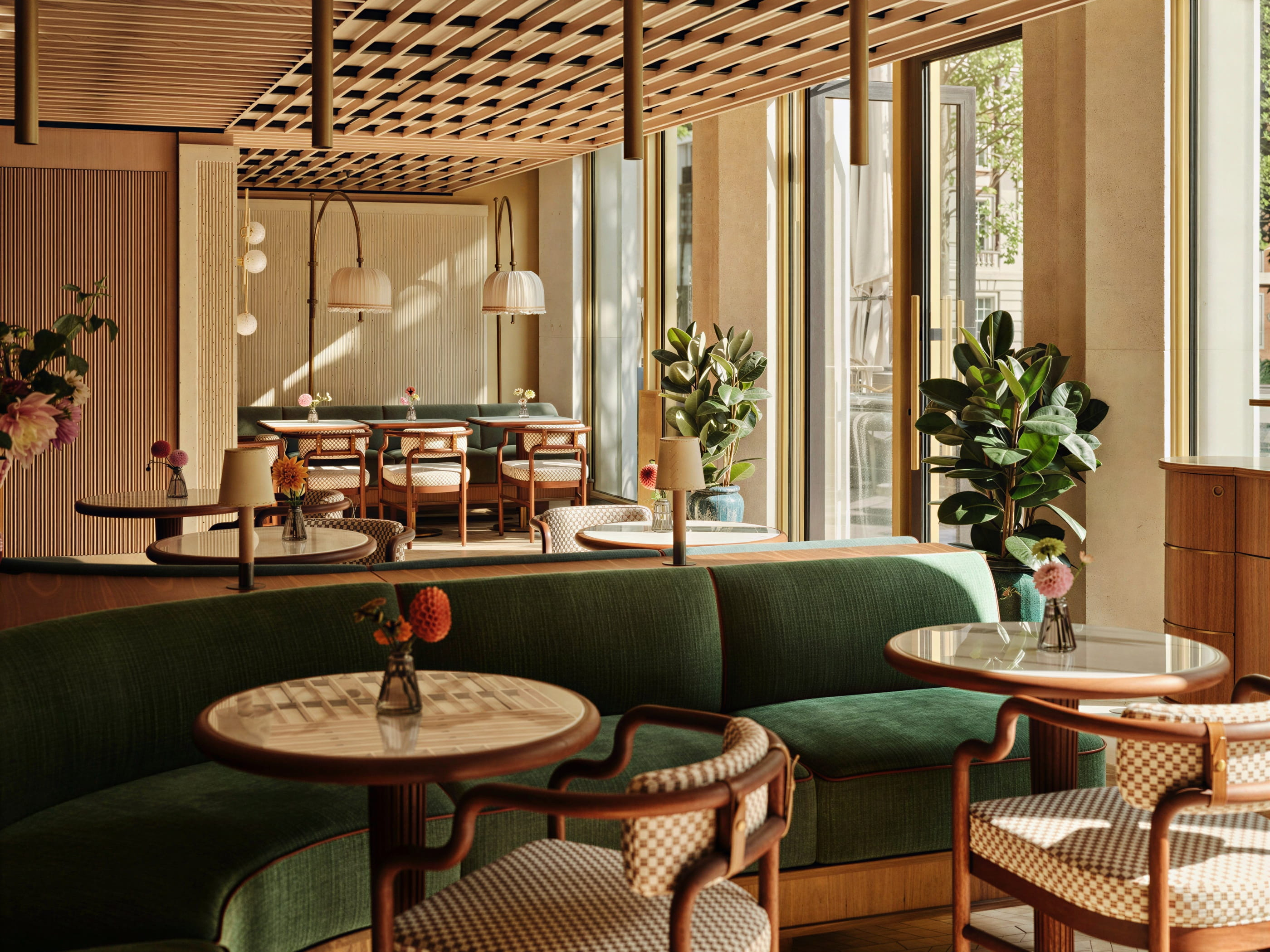 Sunlit cafe interior of Jacqueline at The Rosewood Chancery with green sofas, wooden furniture, and plants. Round tables feature small flower vases. Warm, inviting atmosphere.