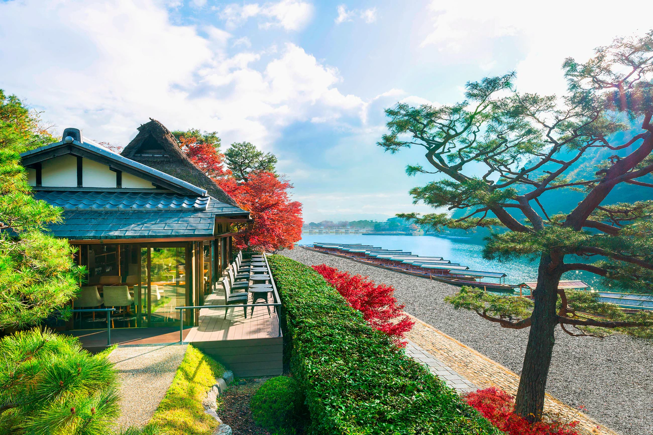 Traditional Japanese house by a lake with vibrant red and green foliage, clear sky, and boats on the water. Peaceful and serene setting.