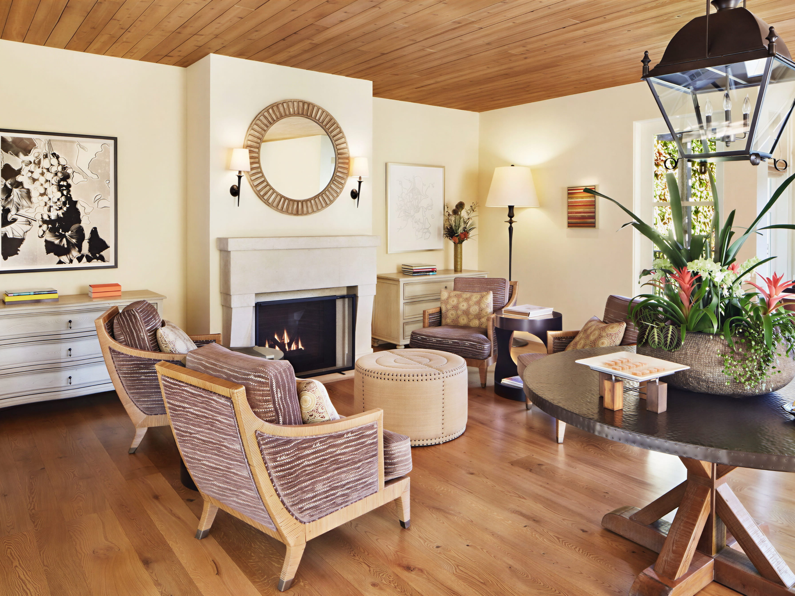 Cozy living room with fireplace, patterned chairs, a round mirror, and a large plant centerpiece on a wooden table. Warm, inviting atmosphere.