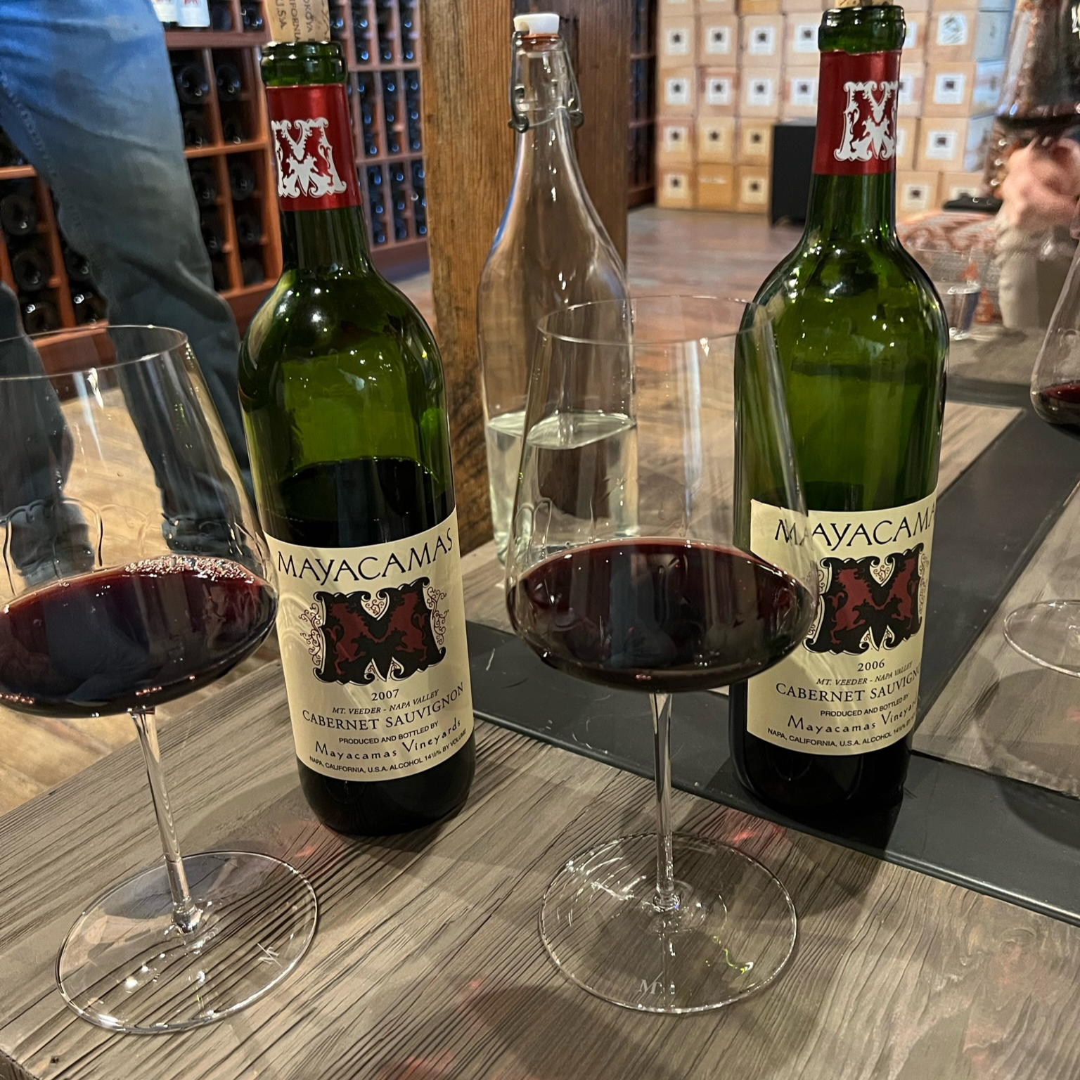 Two Mayacamas wine bottles and filled glasses on a wooden table, in a cozy wine cellar. Visible text: "Mayacamas, Cabernet Sauvignon."