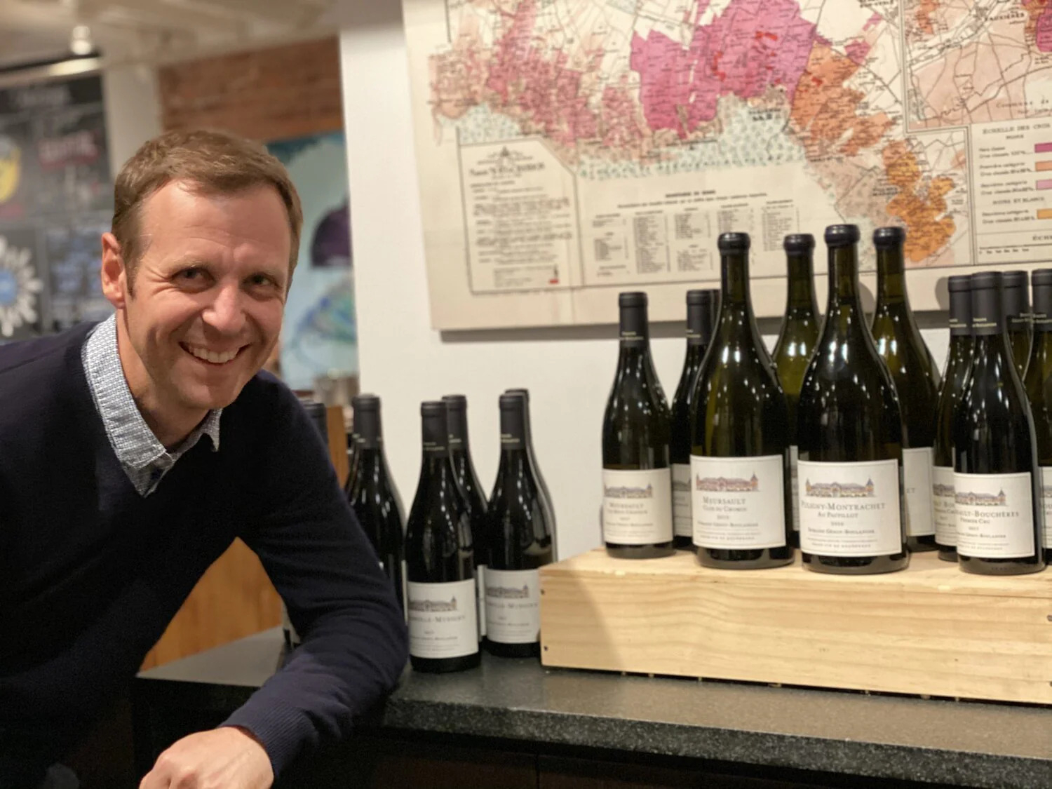 Domaine Génot-Boulanger winemaker Guillaume Lavolée smiles next to his wine bottles on a wooden stand. Background features a colorful map. Warm, inviting atmosphere.