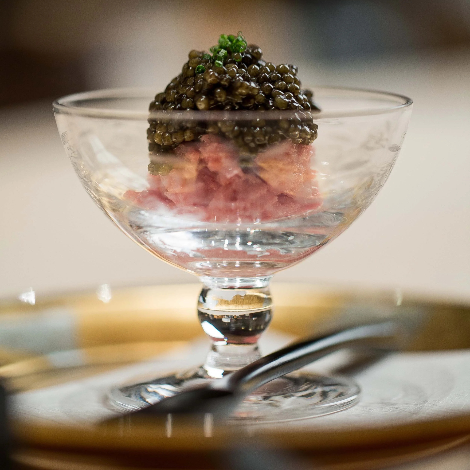 Glass dish with pink meat and black caviar, garnished with herbs. Set on a shiny gold-rimmed plate with a blurred background.