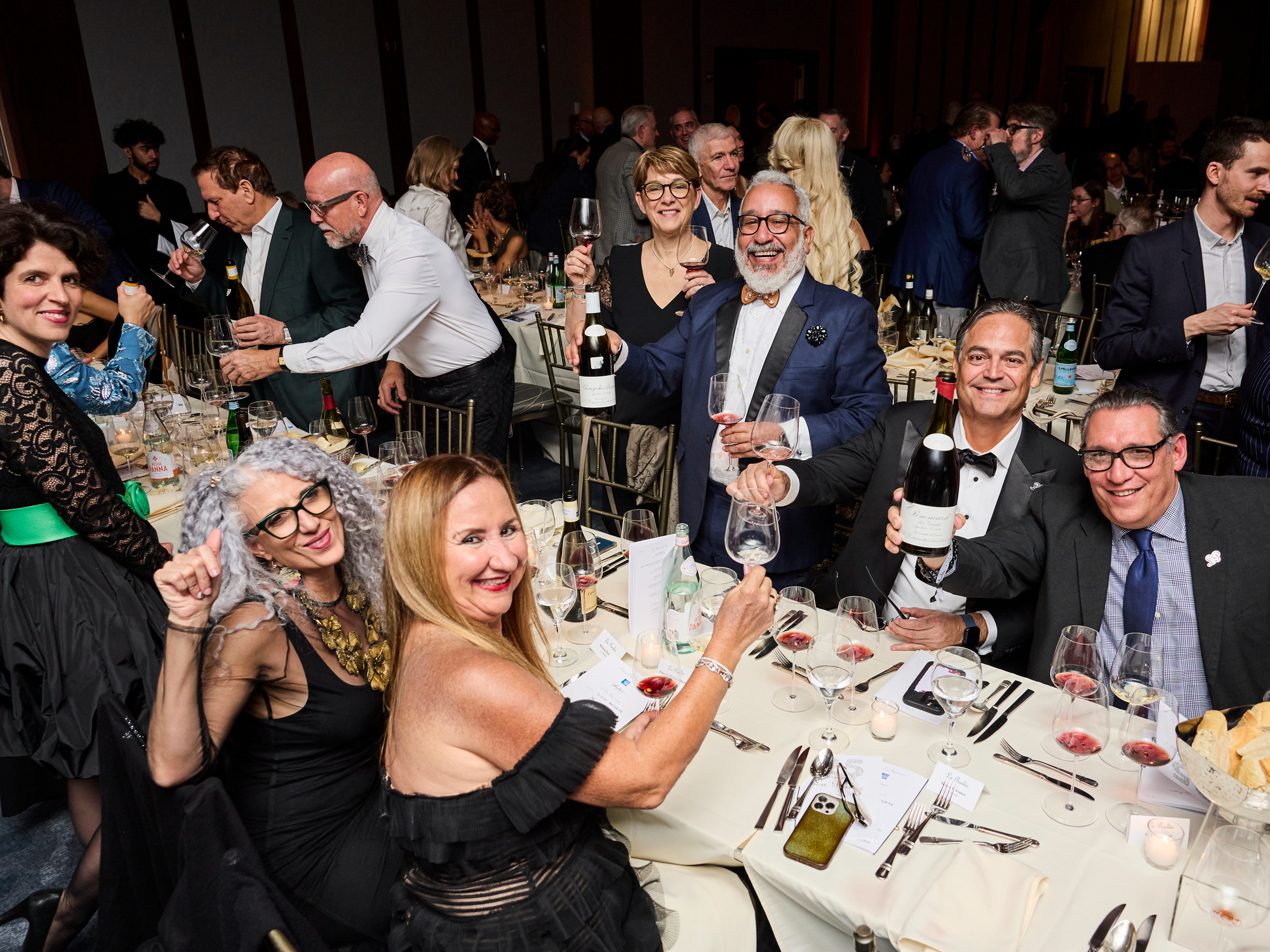 Elegant Revelers at the 2025 Gala Dinner at La Paulée NYC 2025 raise a glass to toast. People in formal wear hold wine glasses and bottles, smiling. Tables set with wine, menus, and candles.