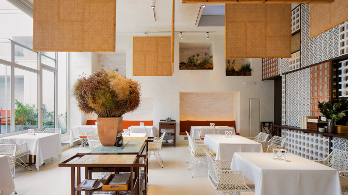 Modern restaurant interior with white tablecloths, wicker chairs, and large potted plant. Geometric wall patterns and soft lighting.