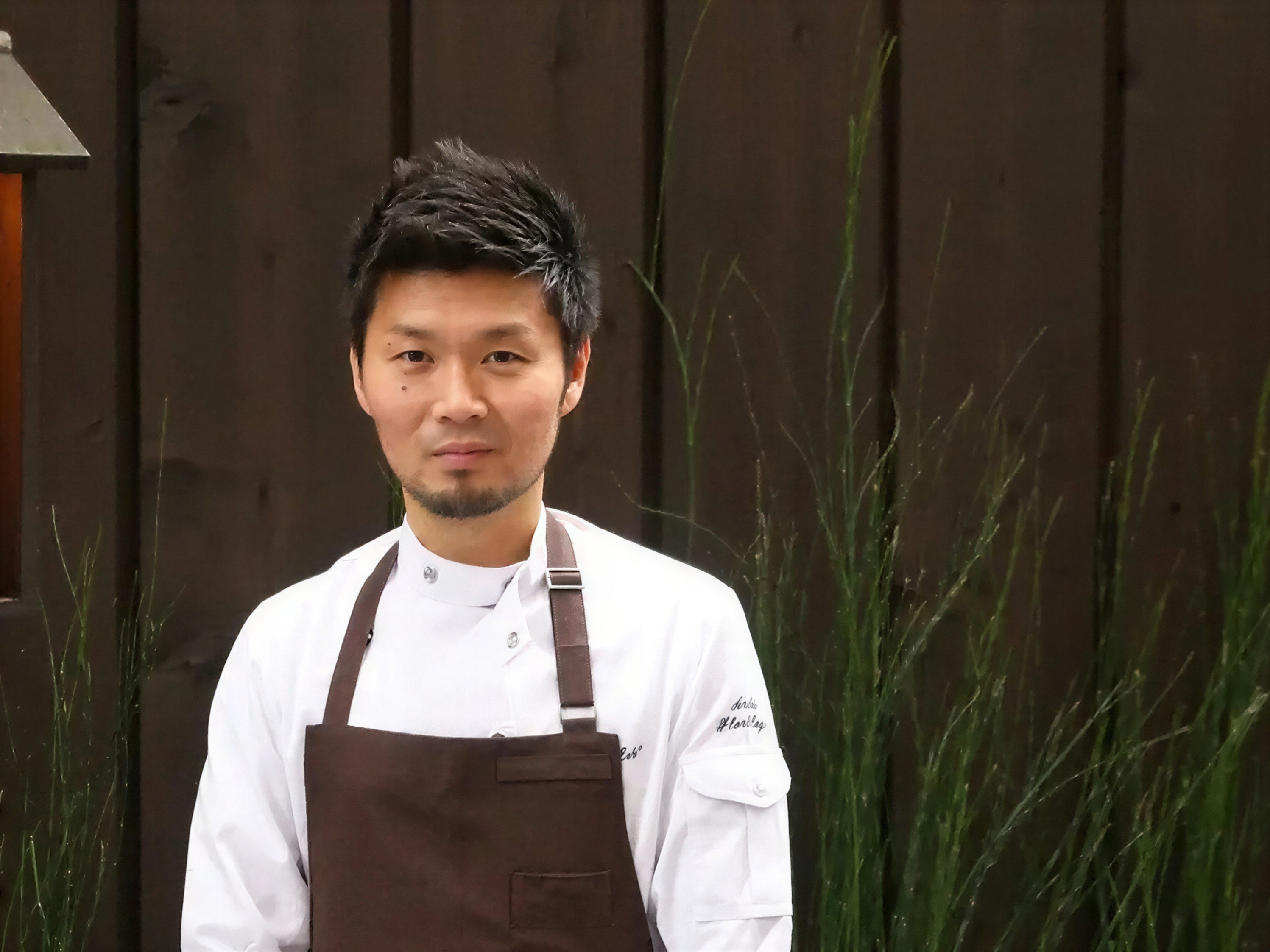 Chef in a white coat and brown apron stands in front of a wooden fence with tall grass. Calm expression, neutral background.