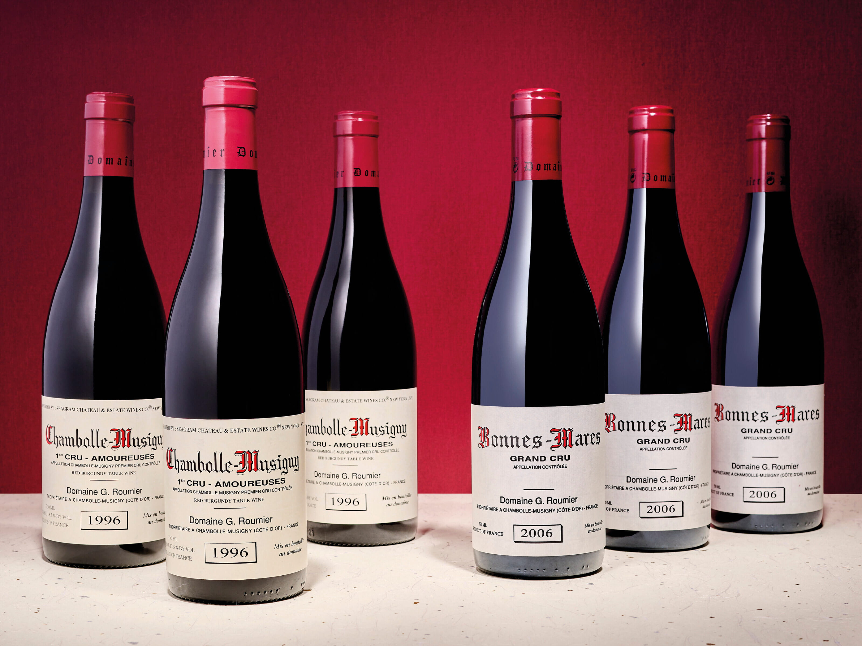 Six wine bottles of 1996 Domaine Georges Roumier Chambolle-Musigny Les Amoureuses and 2006 Bonnes Mares with red labels. They stand on a light surface against a red background.