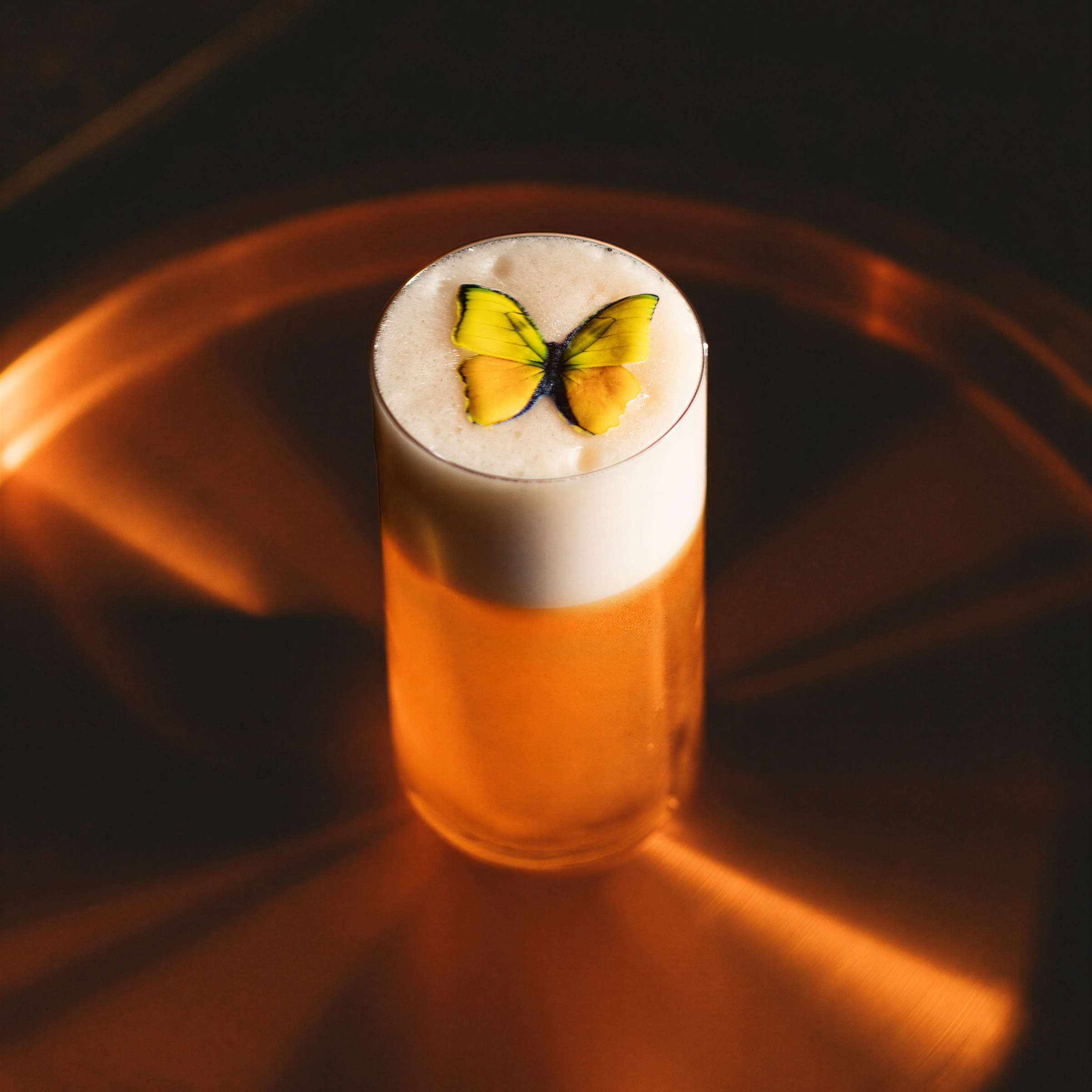 A tall glass of foamy beer with a yellow butterfly decoration on top, set against a warm, diffused copper-toned background.