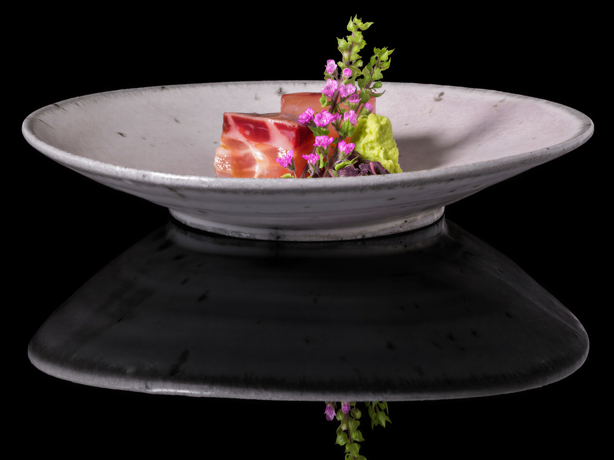 Gourmet dish on a white plate with slices of meat, green garnish, and pink flowers, set against a black background, reflecting below.