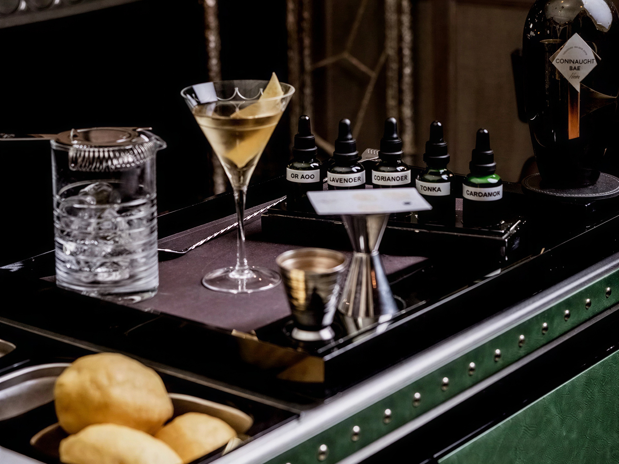 Cocktail station with a martini, ice-filled mixing glass, various aromatic bottles labeled (Lavender, Coriander), and lemons on a green counter.