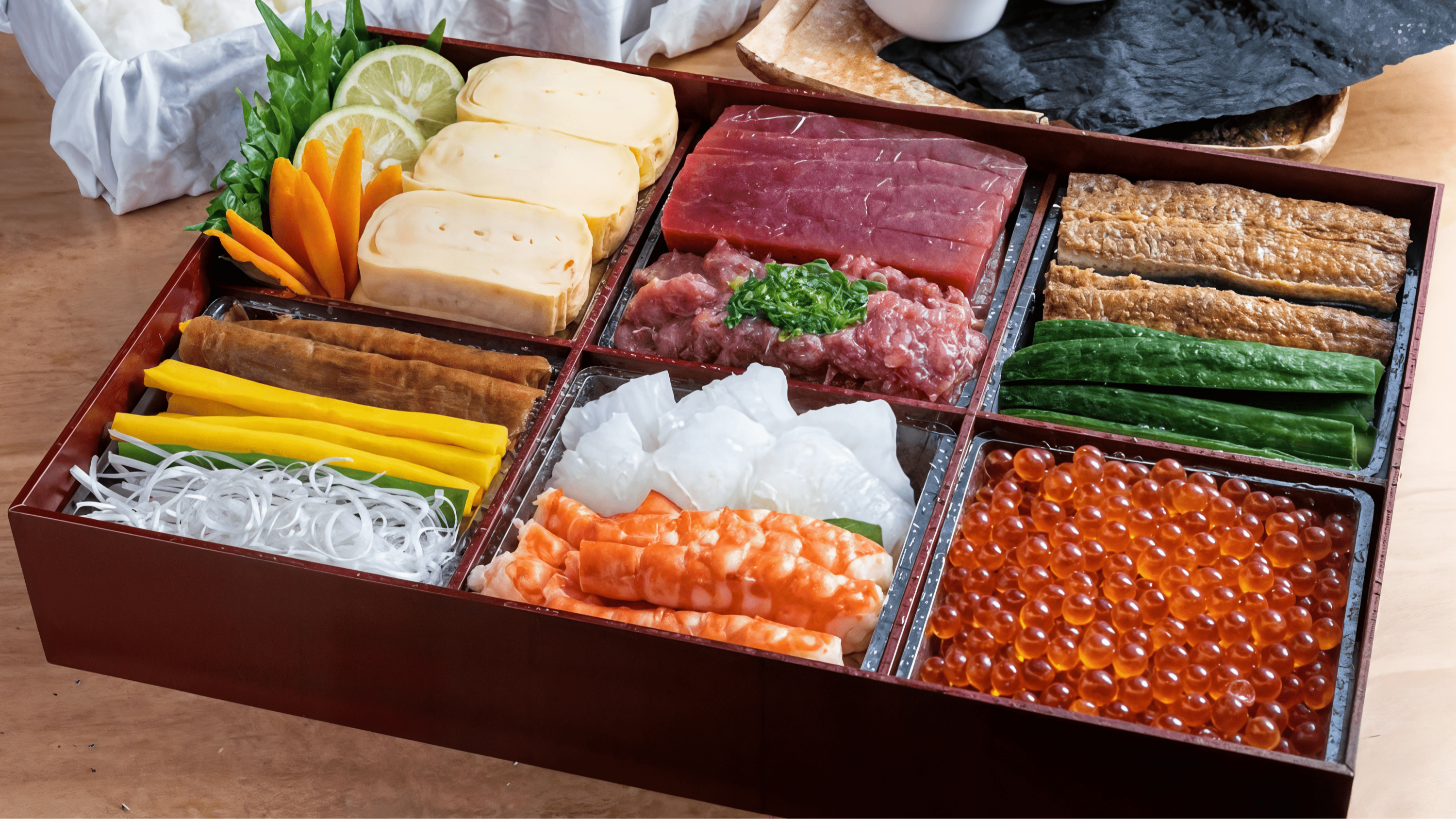 A wooden box with assorted sushi ingredients: egg, tuna, cucumber, radish, shrimp, and orange roe. Set on a light wooden table.