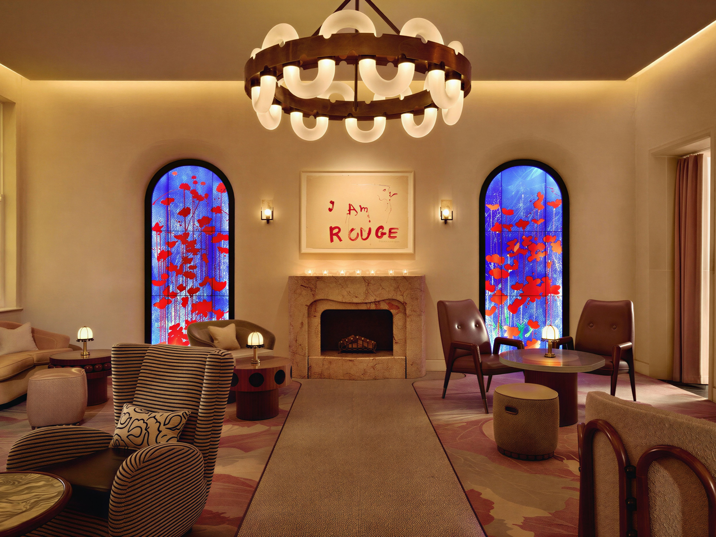 Cozy lounge with striped chairs, warm lighting, and stained glass windows. Art reads "I AM ROUGE" above a stone fireplace. Elegant decor.
