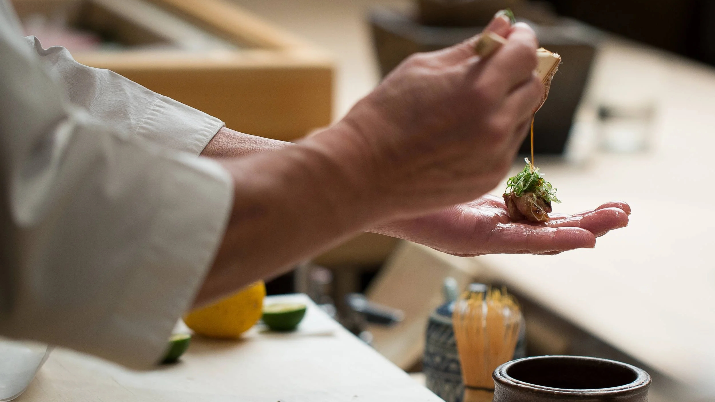 Chef drizzles sauce over sushi in hand, set against a blurred kitchen background. Close-up with neutral tones and a focused mood.