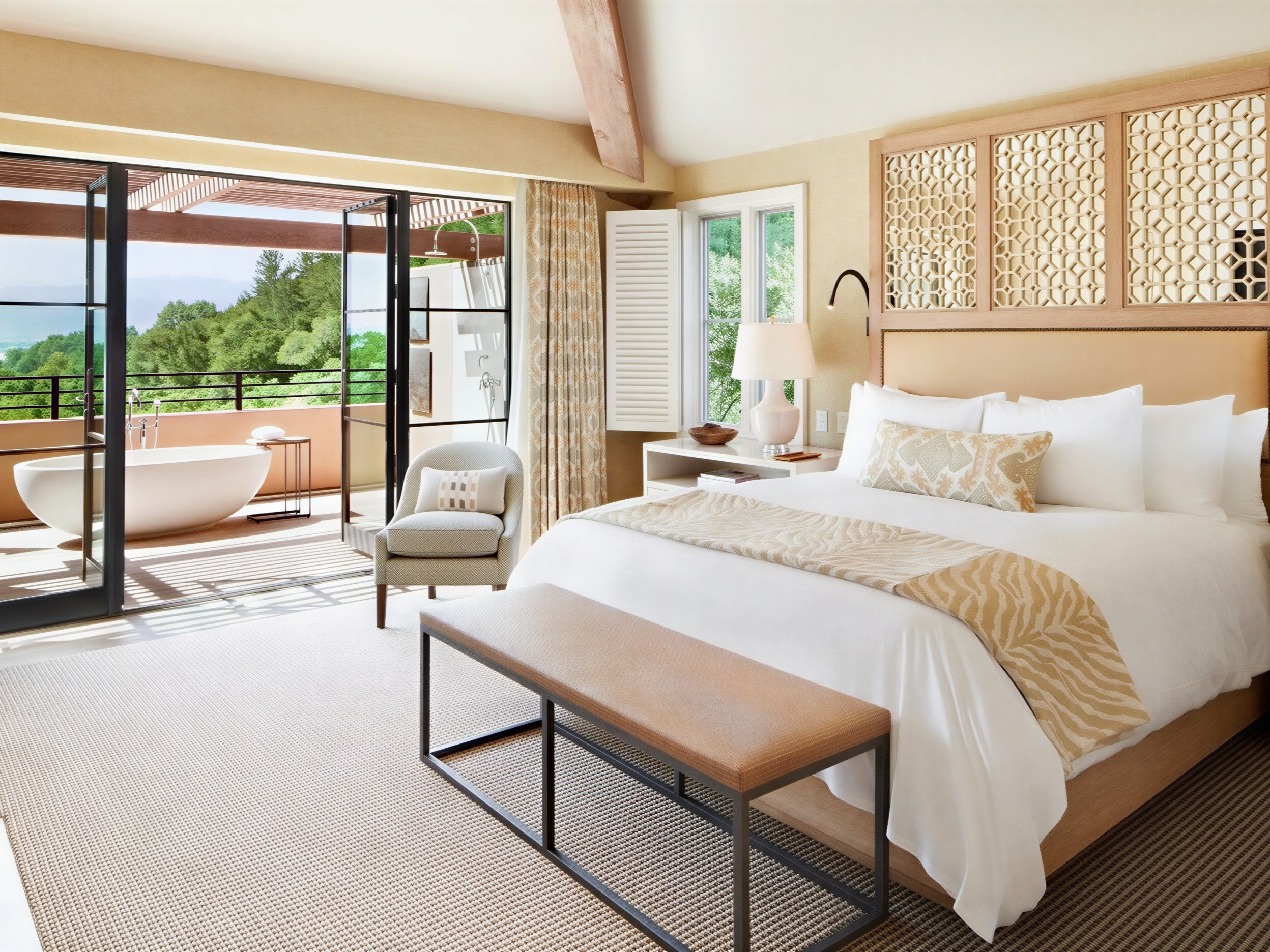 Elegant bedroom with a large bed, patterned throw, and open doors to a balcony with a freestanding tub. Neutral tones, natural light.