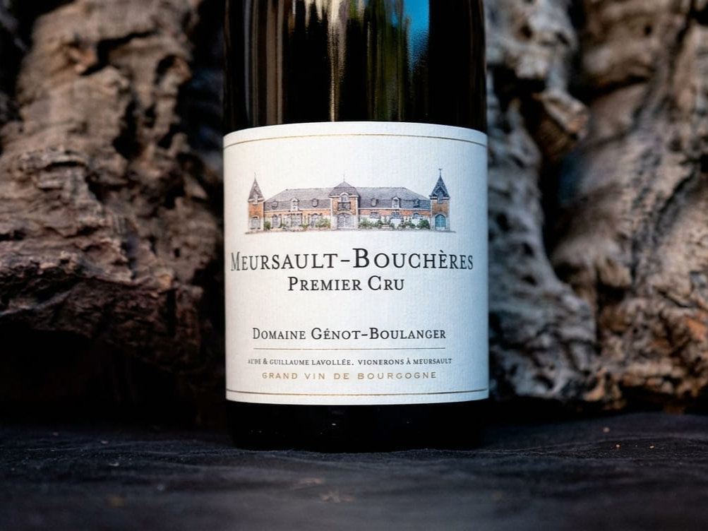 Wine bottle of Domaine Génot-Boulanger Meursault-Bouchères Premier Cru on dark surface, with a textured, rustic wood background. Label features estate image.