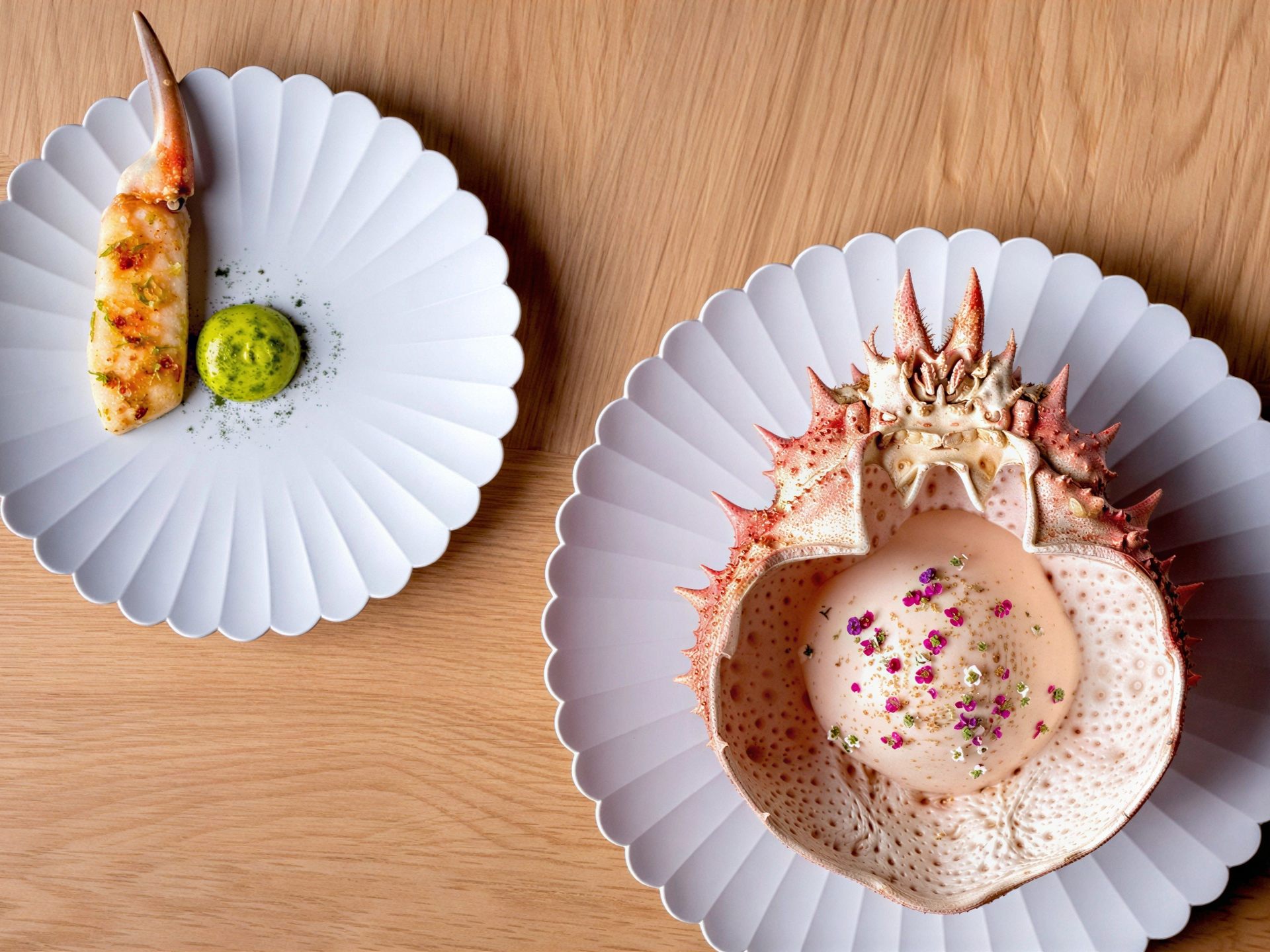 Crab claw and crustacean cap artfully plated on white scalloped dishes atop a wooden table. Garnishes add pops of green and pink.