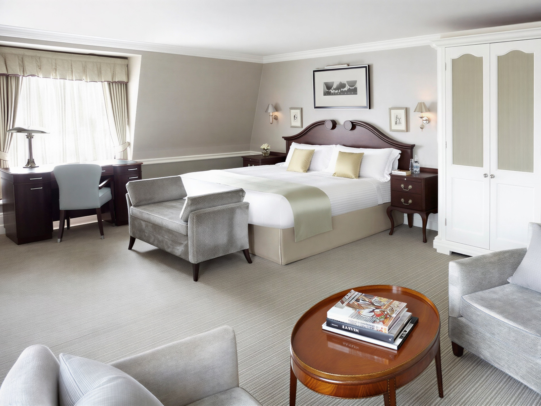 Elegant hotel room with a large bed, soft gray chairs, wooden desk, and side tables. Neutral tones, bright window, calm ambiance.