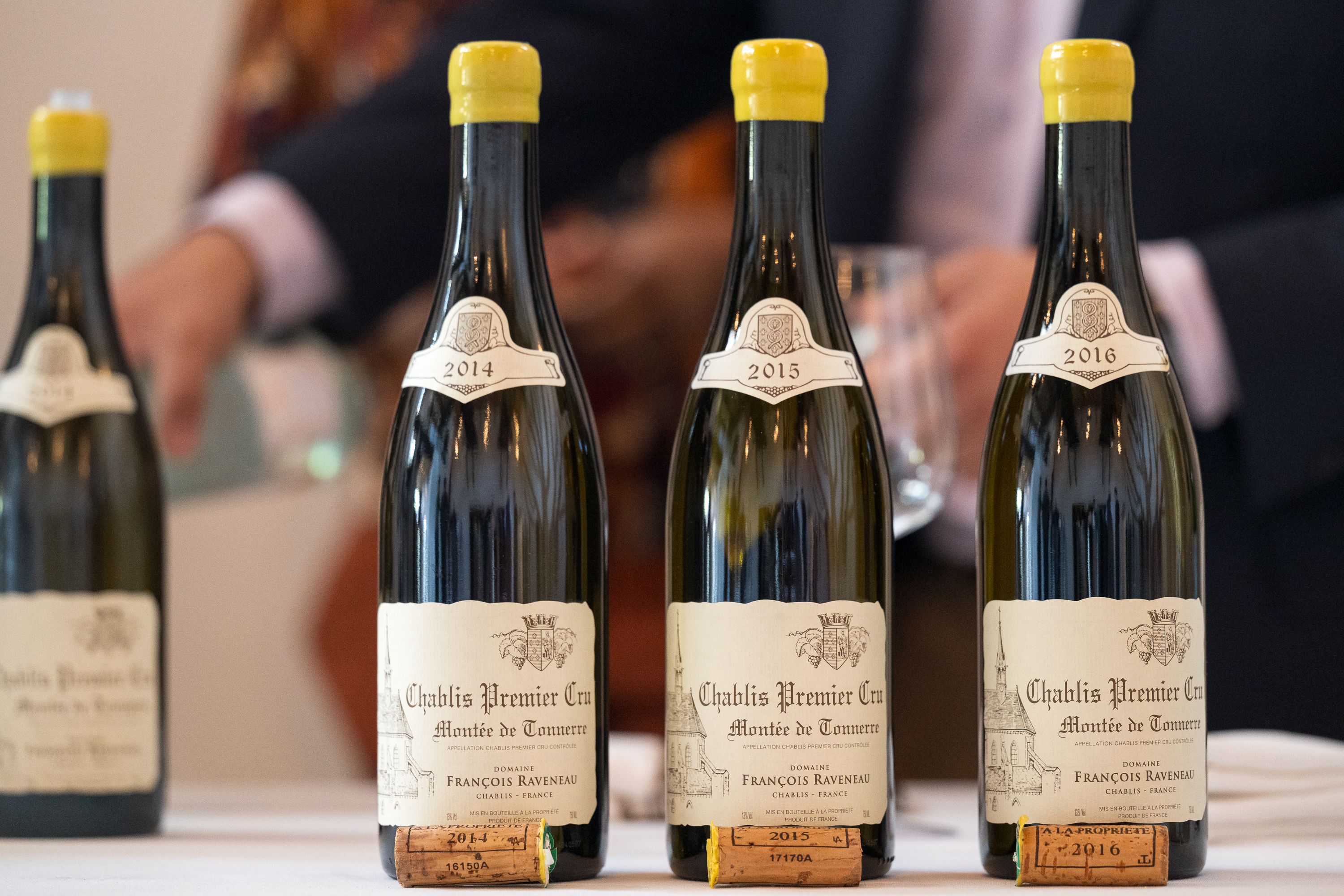 Three bottles of Chablis Premier Cru (2014, 2015, 2016) on a table with corks. Blurred figures and wine glass in background.