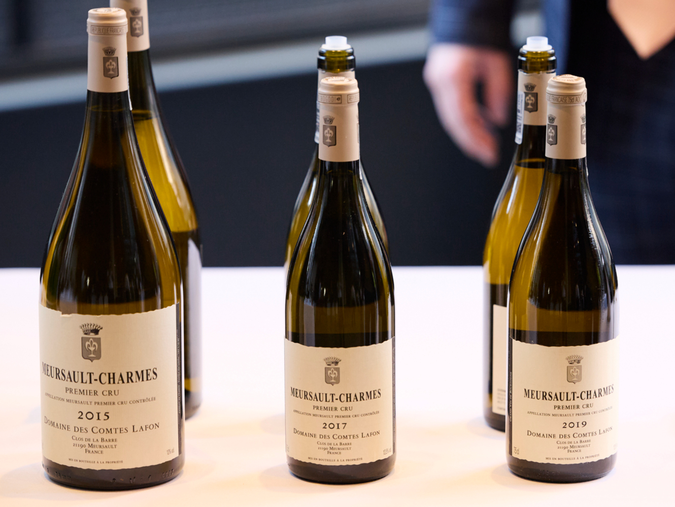 3 vintage vertical of 2015, 2017 and 2019 Domaine des Comtes Lafon Meursault-Charmes on a table, alongside a San Pellegrino bottle. A person in a suit stands nearby, holding a glass.