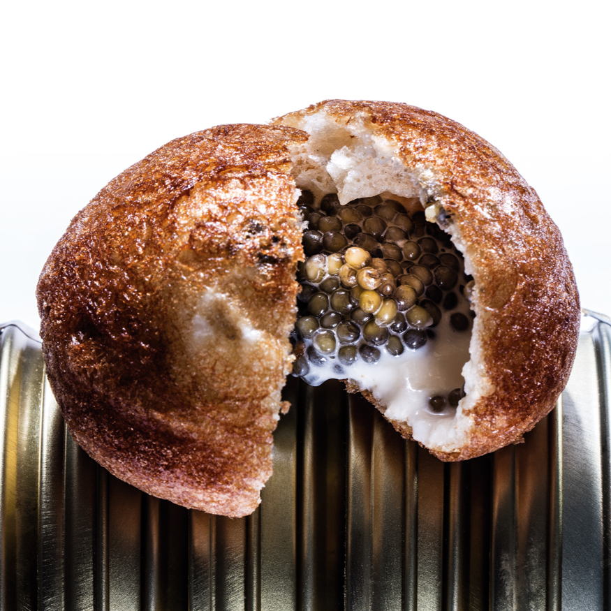 A crispy bread roll, cut open to reveal caviar and cream filling, sits atop a corrugated tin. White background enhances focus.