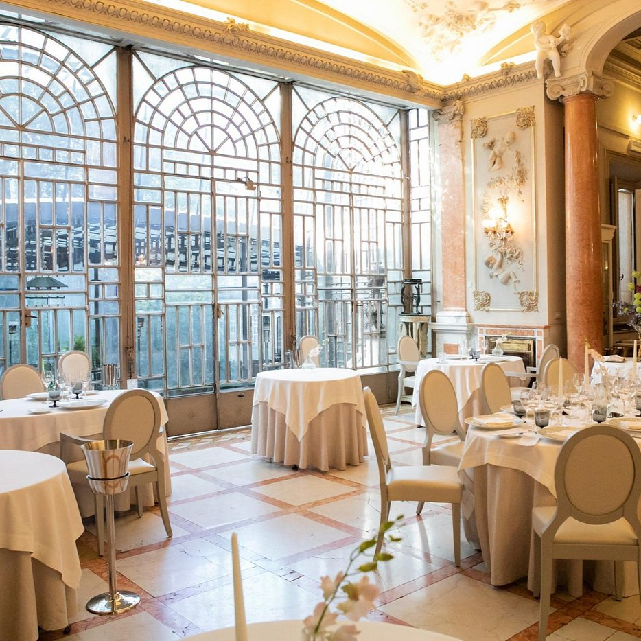 Elegant dining room with round tables, white tablecloths, and beige chairs. Large ornate windows and columns. Bright and sophisticated ambiance.