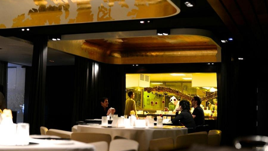 Dimly lit restaurant interior, patrons at tables, visible open kitchen with chefs inside. Warm tones, elegant decor, relaxed ambiance.