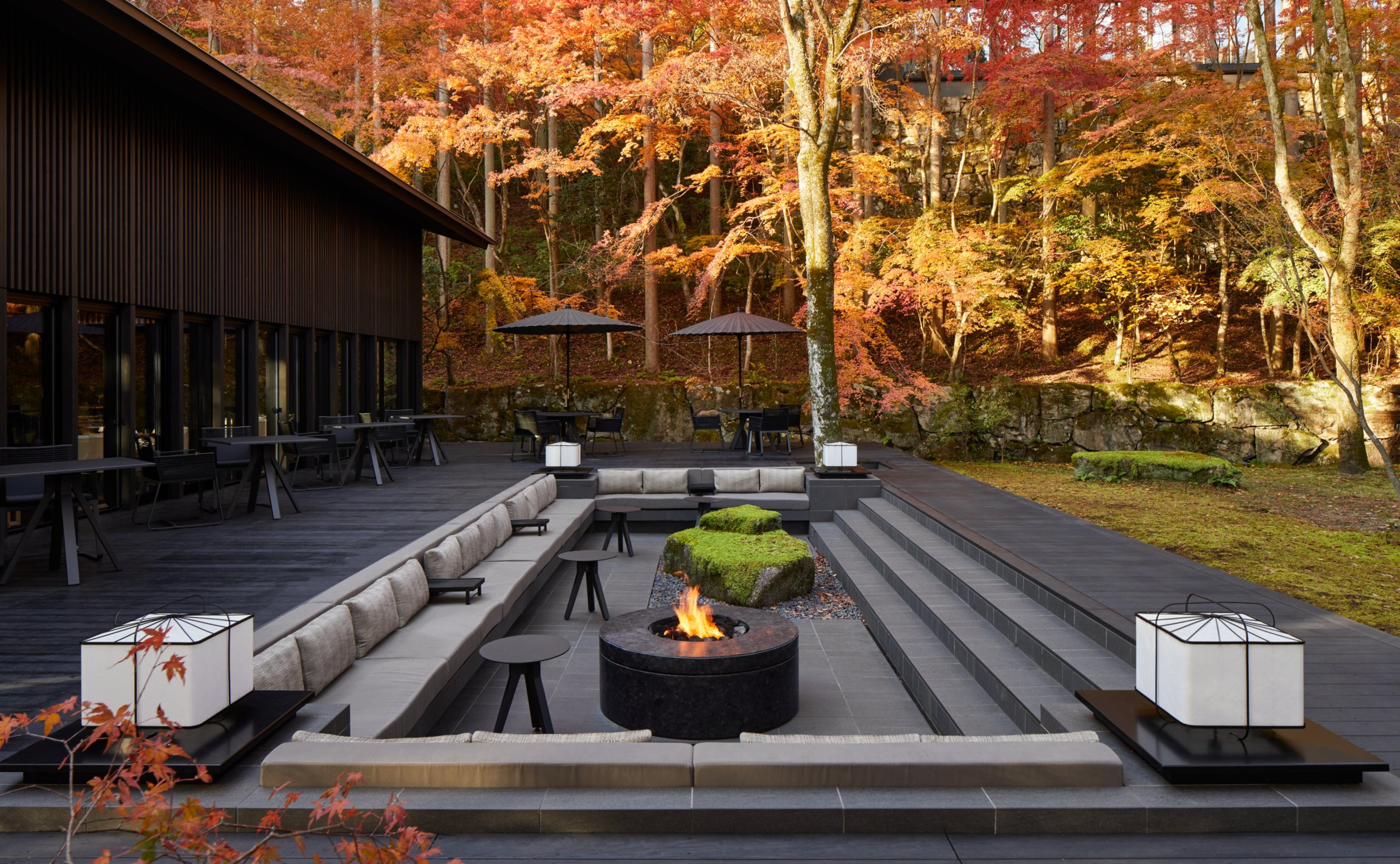 Outdoor seating area with a central fire pit, surrounded by autumn trees. Modern furniture and lamps, creating a cozy, serene atmosphere.