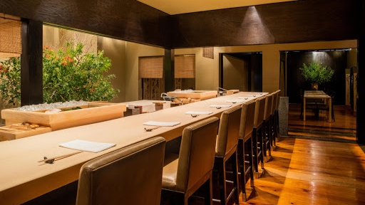 Elegant sushi bar with brown chairs, light wood counter set with napkins and chopsticks, and warm lighting. Greenery decorates the background.