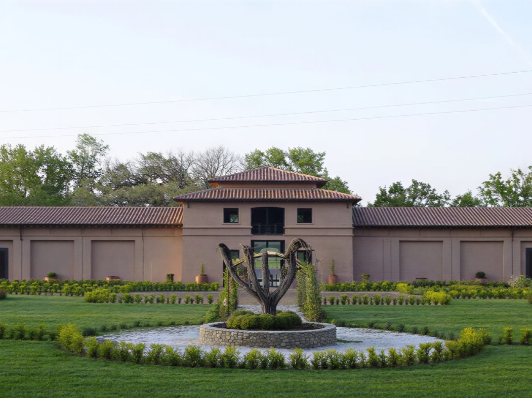 Elegant building of Tenuta San Guido estate with terracotta roof, surrounded by lush greenery. Central sculpture in front, clear sky, calm and serene setting.